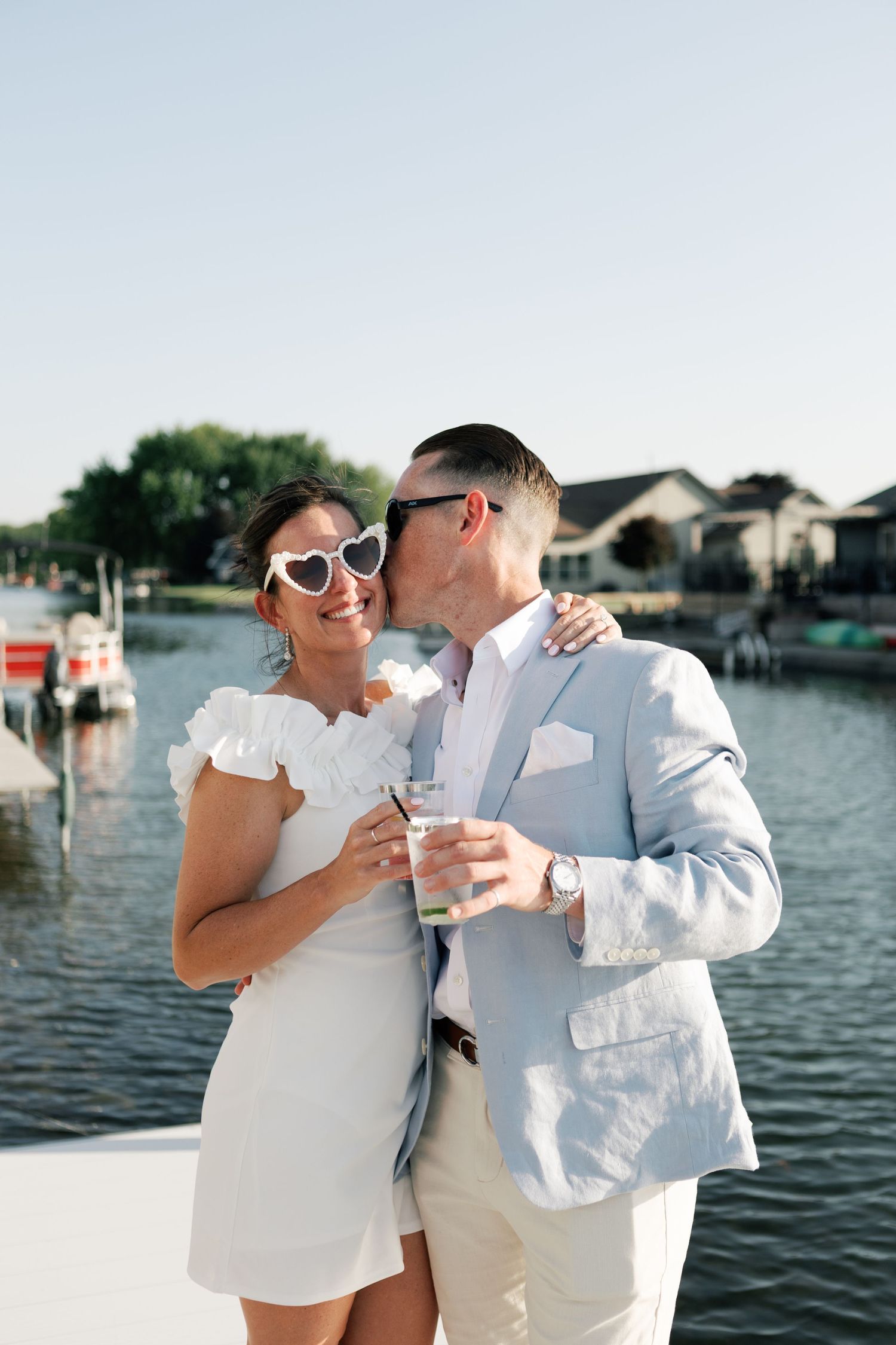 Couple in white and beige outfits share drinks by waterfront during sunset.