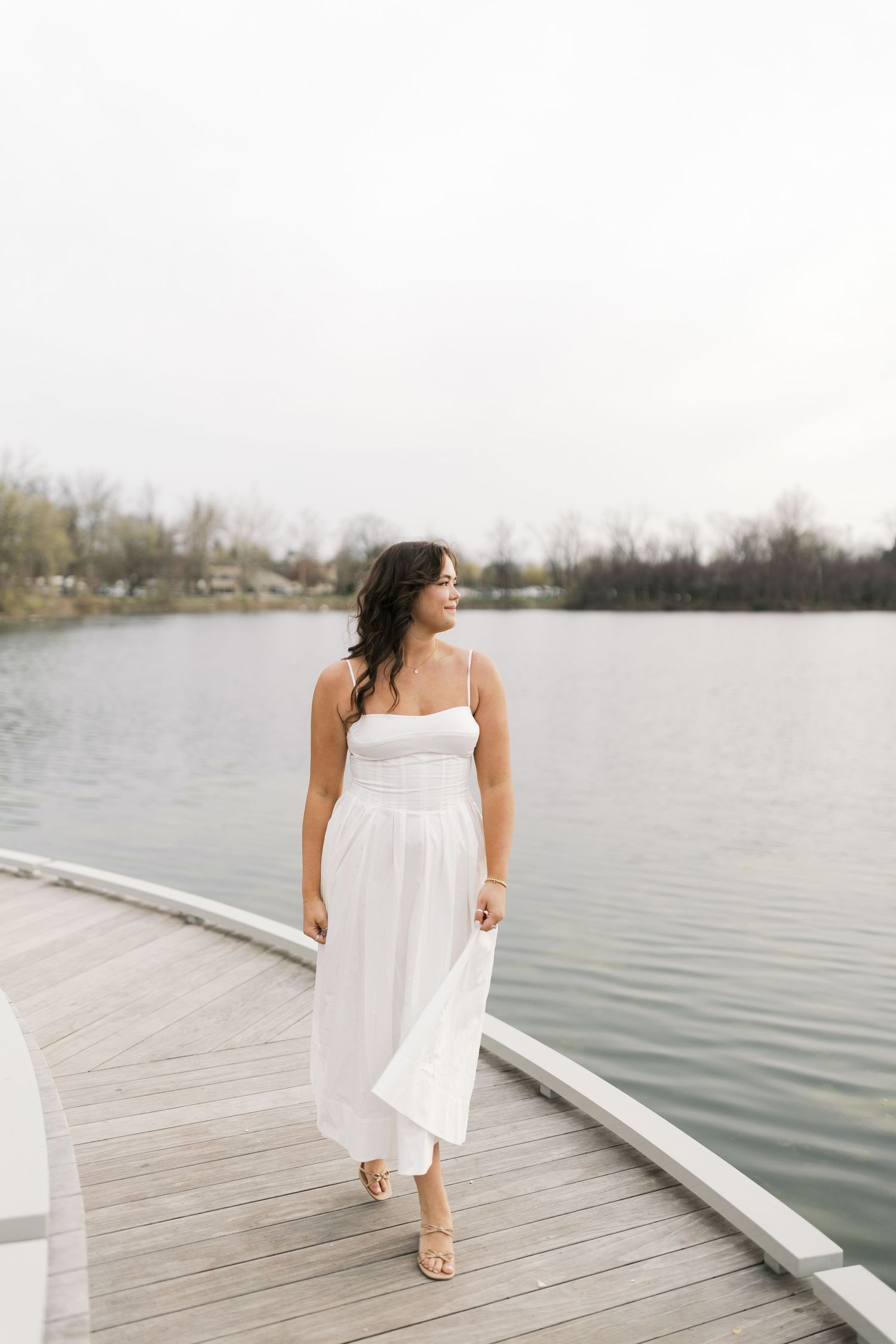 A woman in a flowing white sundress walks along a wooden dock by a serene lake on an overcast day.