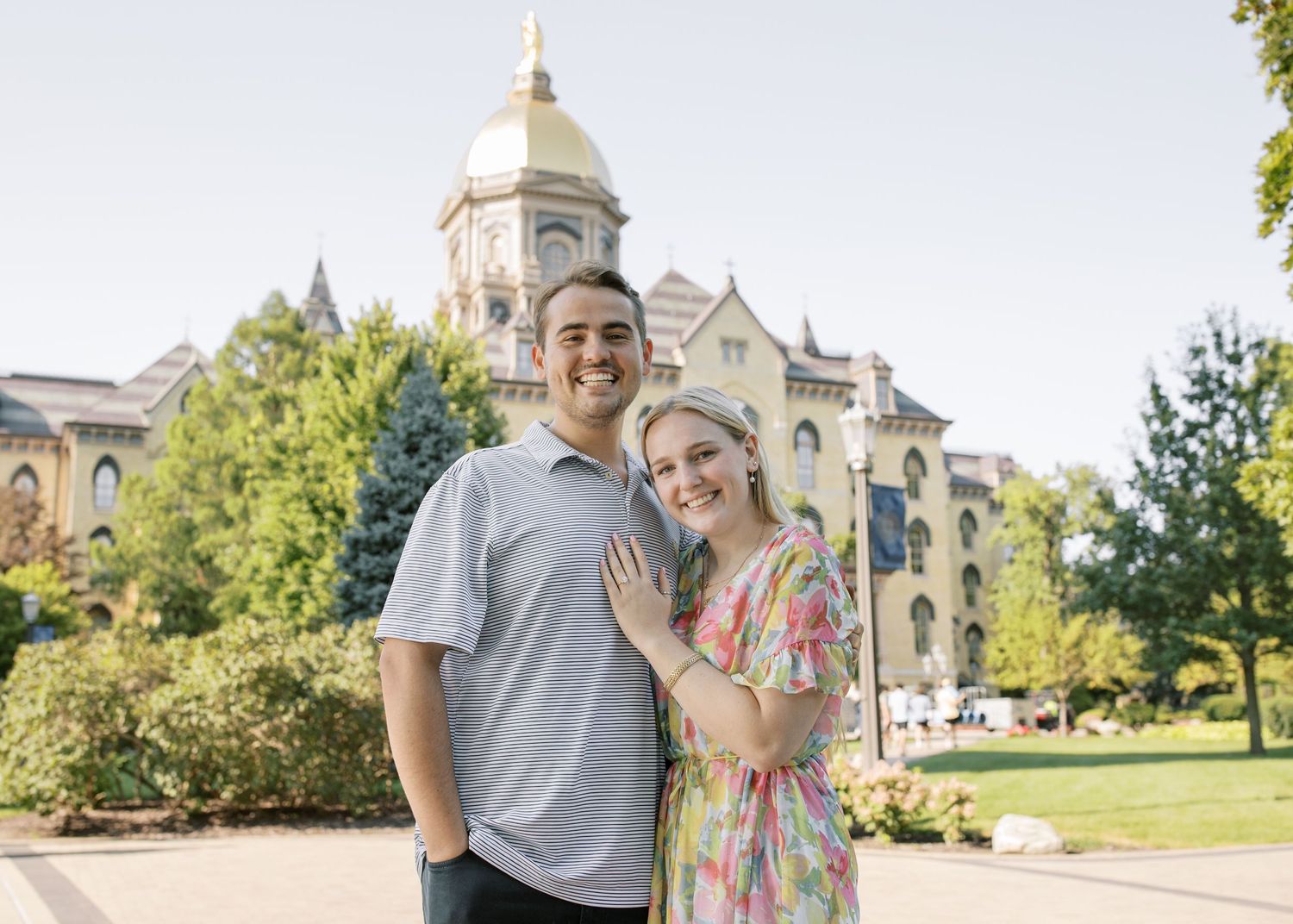 A couple poses together in front of a grand collegiate building with a golden dome on a sunny day.