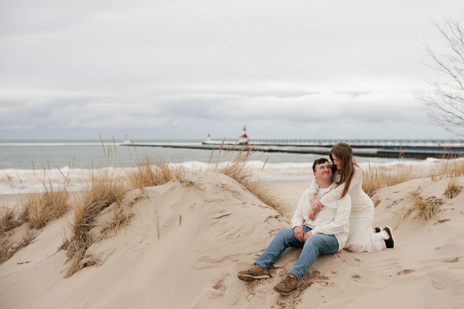 Couple sitting on sand dunes at a beach with a lighthouse pier in the background on a cloudy day.