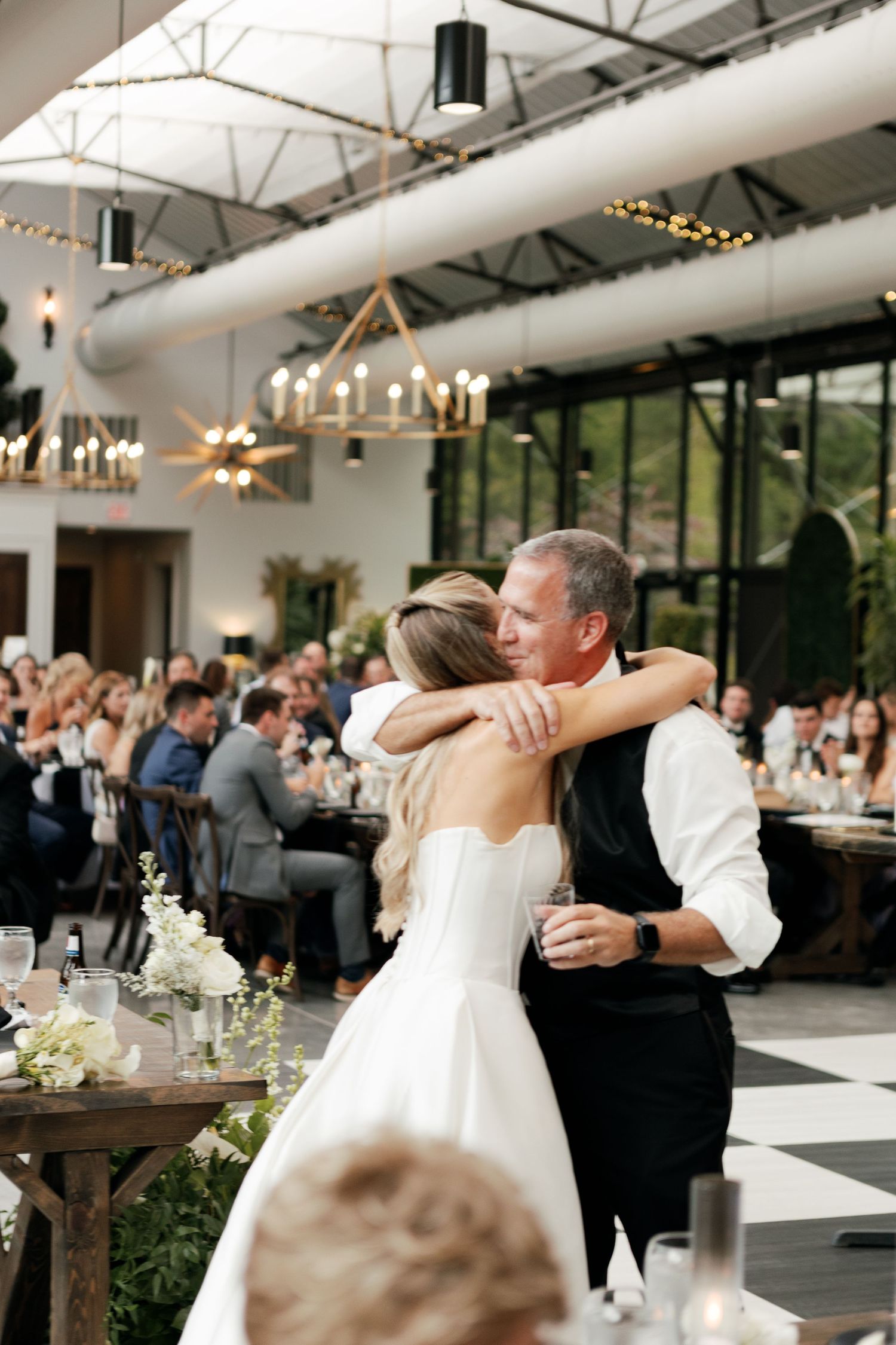 Couple dancing together at an elegant wedding reception with chandeliers and rustic wooden tables.