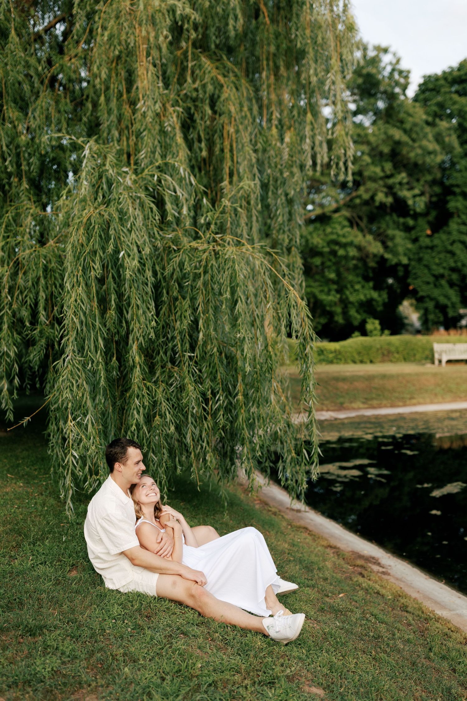Two people rest together on a sloping lawn beside a pond with drooping willow branches overhead.