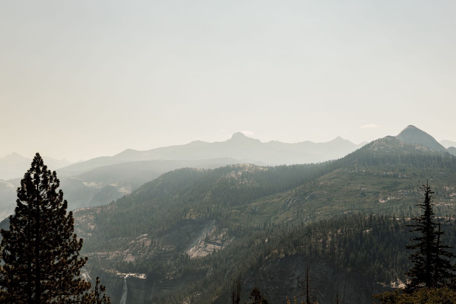 Panoramic mountain landscape with misty peaks and evergreen trees silhouetted against hazy sky.