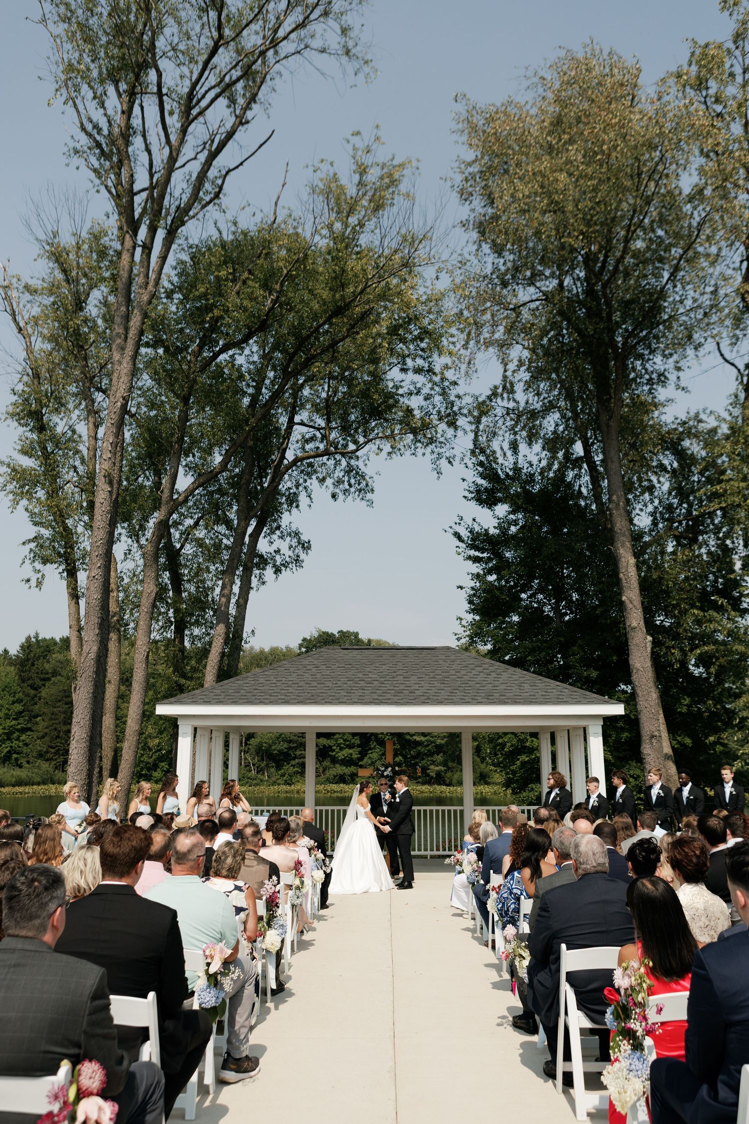 An outdoor wedding ceremony setup with white chairs and guests seated under tall trees.