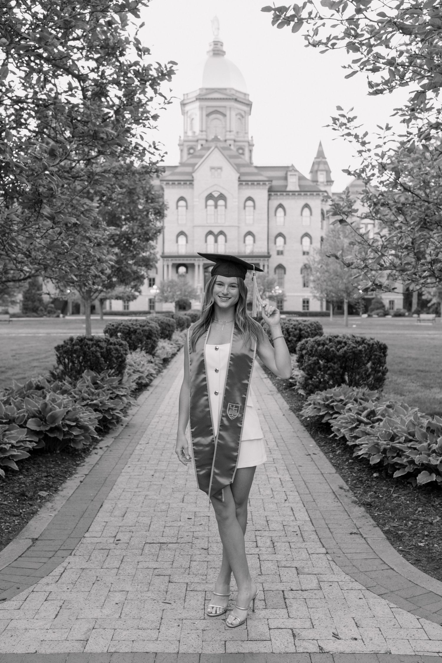 Graduate poses for progression photos on brick path leading to historic Gothic building.