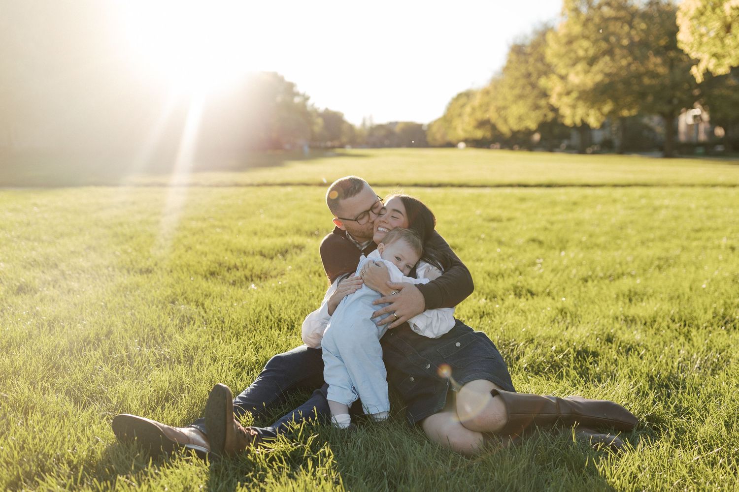 Couple sitting on green grass in sunlit park, kissing baby between them during golden hour outdoor family photo.
