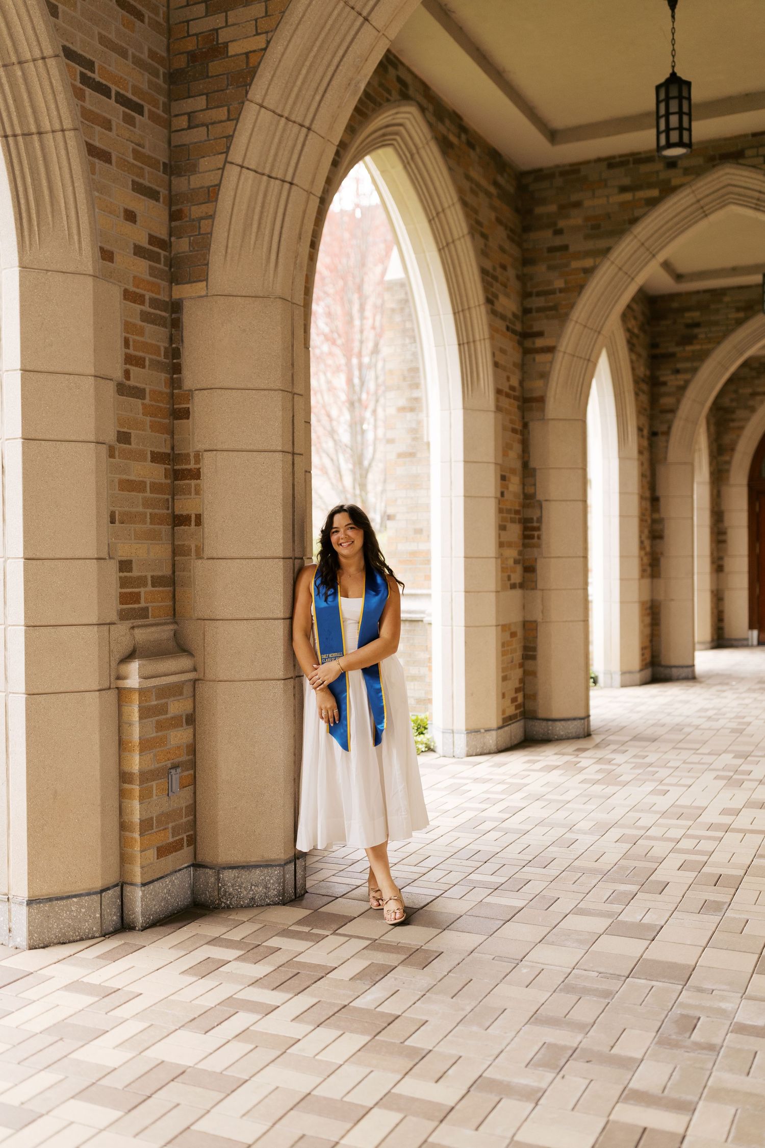 A long arched hallway with stone columns and brick walls features a figure standing and leaning against a pillar.