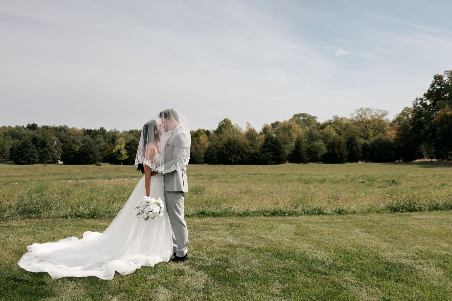 Couple stands together in open field on their wedding day with flowing white dress and veil.
