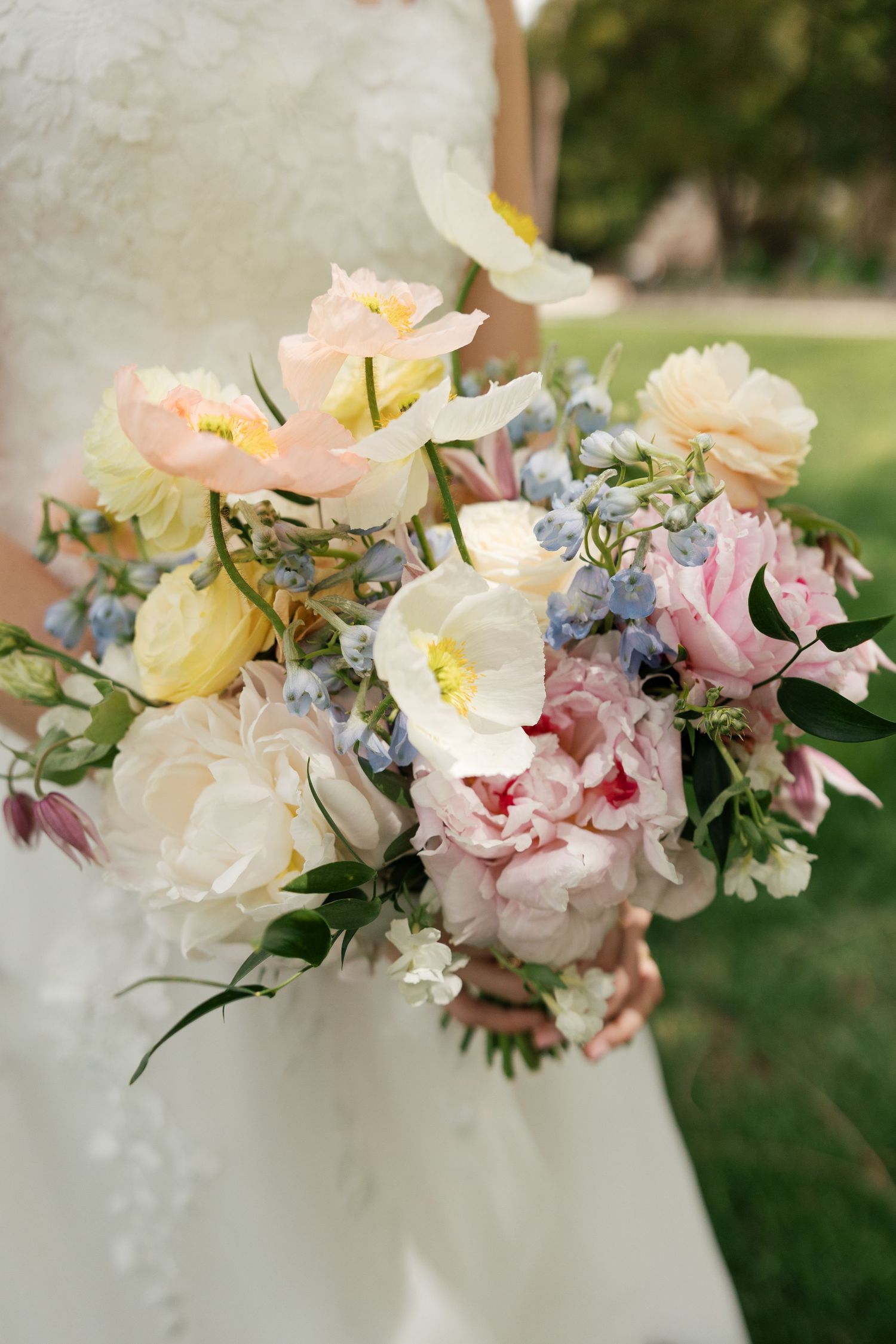 Bride holding a lush wedding bouquet with pink peonies, white tulips, yellow roses, and blue forget-me-nots outdoors.