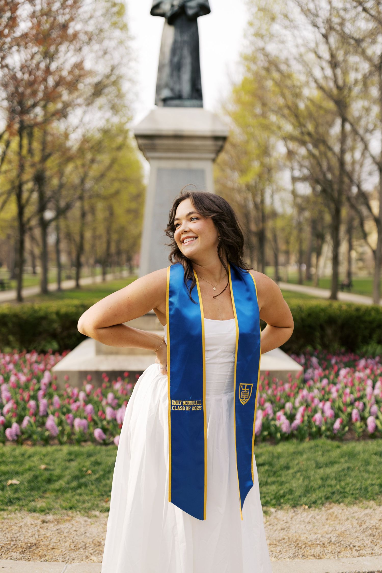 Graduate in a blue and gold honor stole poses for photos in front of spring flowers and trees on campus.