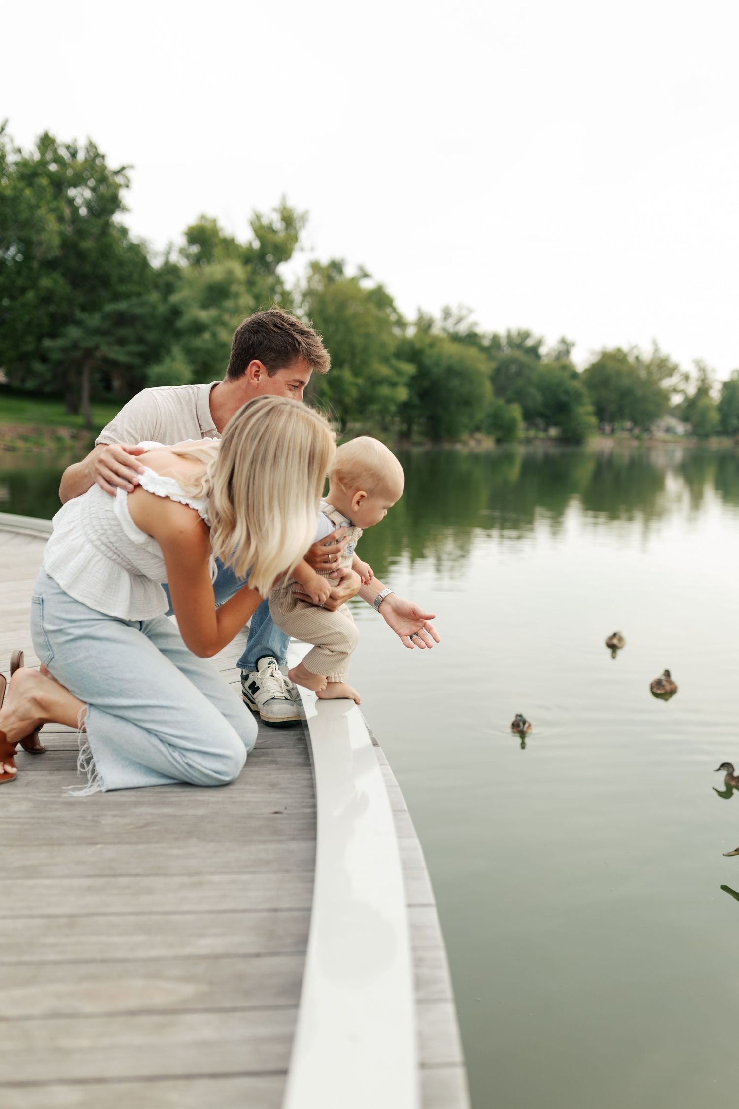 People leaning over dock railing to look at ducks swimming in the lake below.