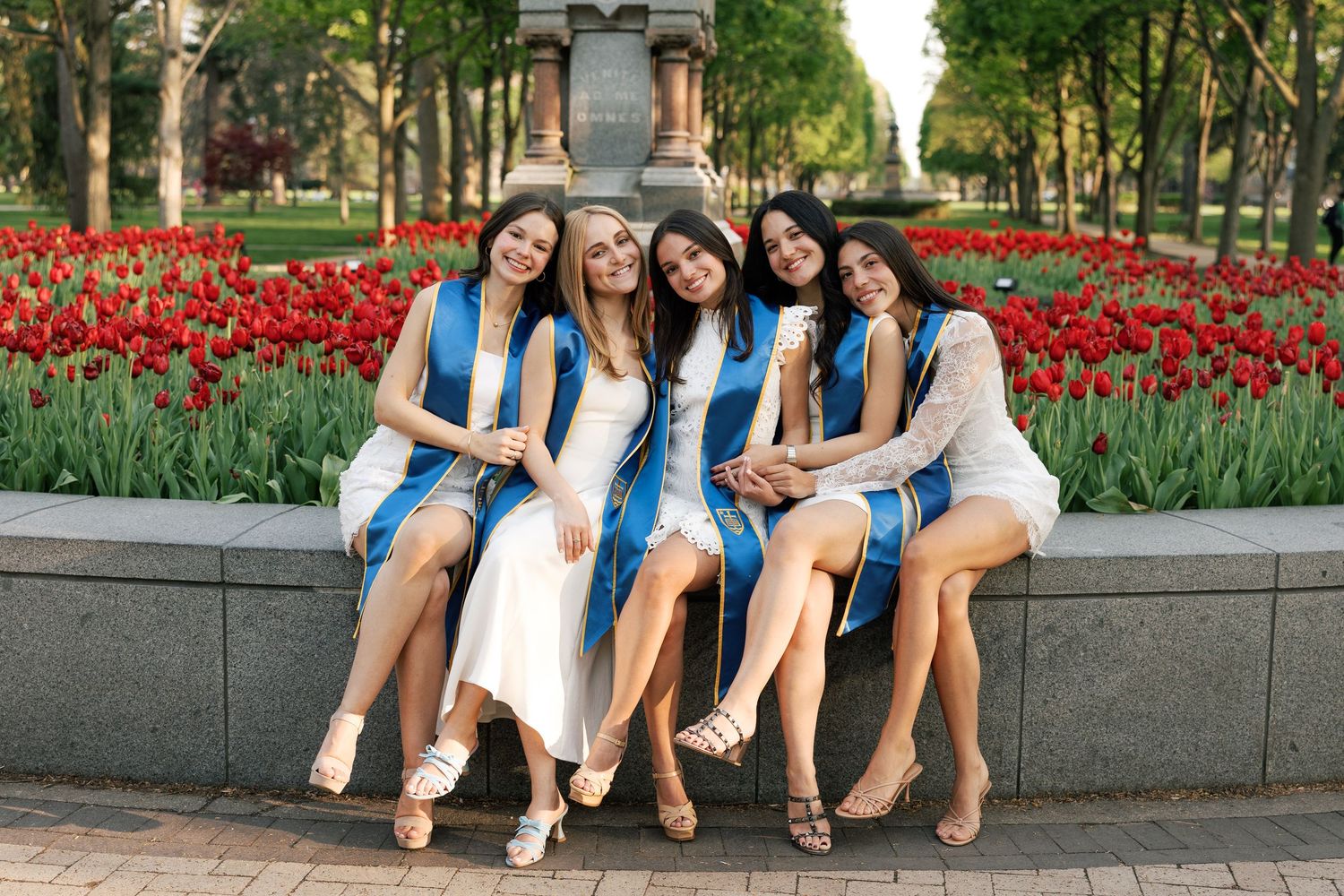 Five young women in white dresses and blue graduation sashes sit together outdoors near red tulips, smiling joyfully.