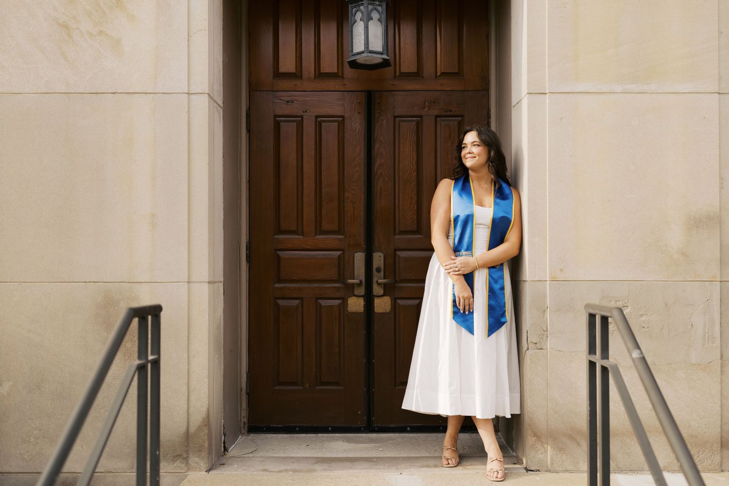 A woman in a blue and white dress poses for graduation photos in front of an elegant stone building entrance.
