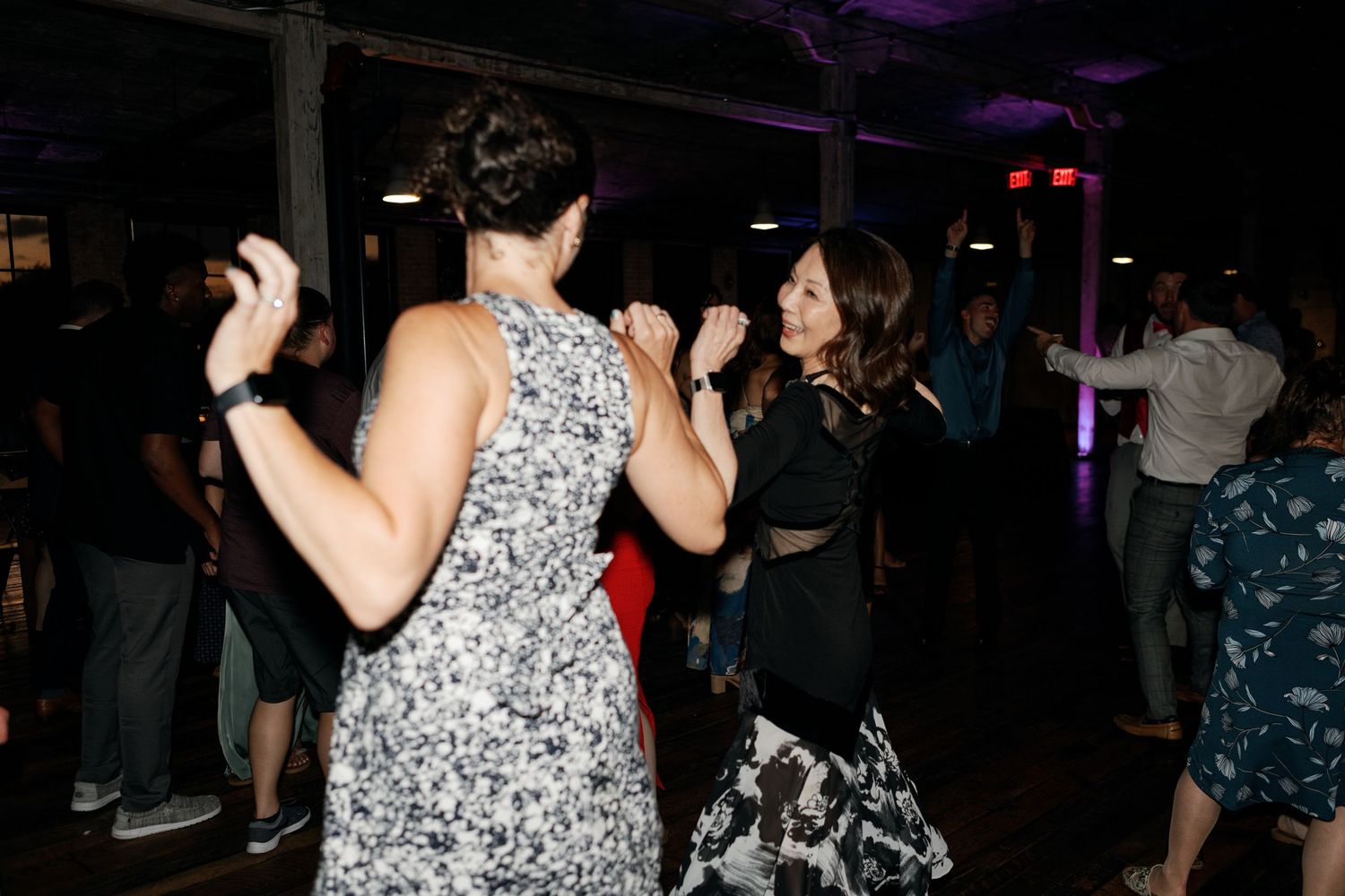 People dancing and celebrating at a dimly lit indoor reception with purple lighting.