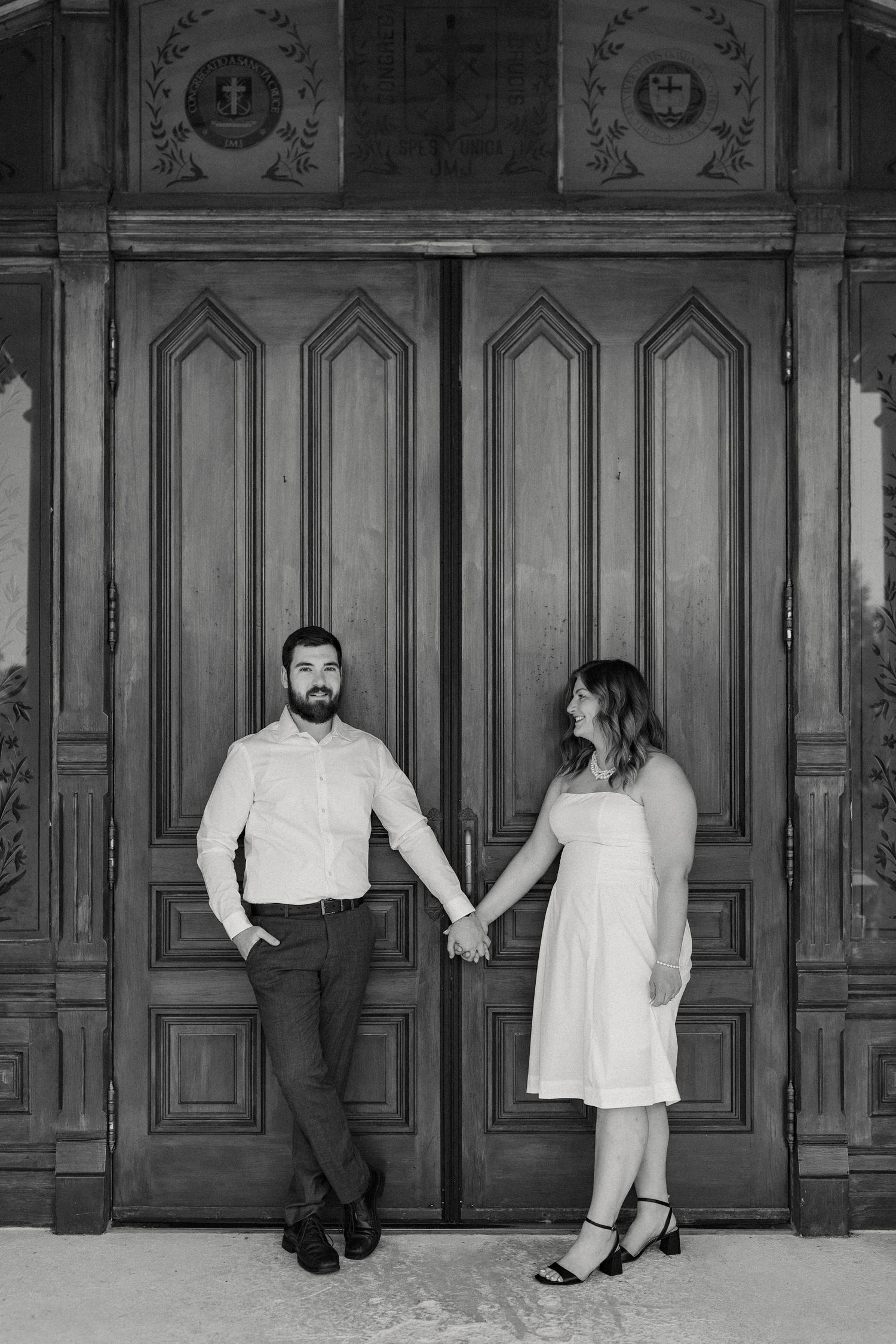 A couple poses playfully in front of ornate wooden doors in a vintage-style black and white photo.