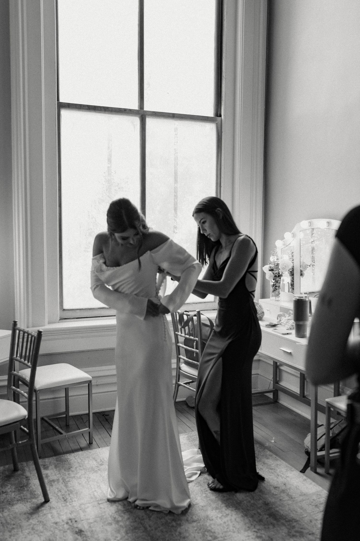 Wedding preparation moment in black and white by large window with elegant dress details.