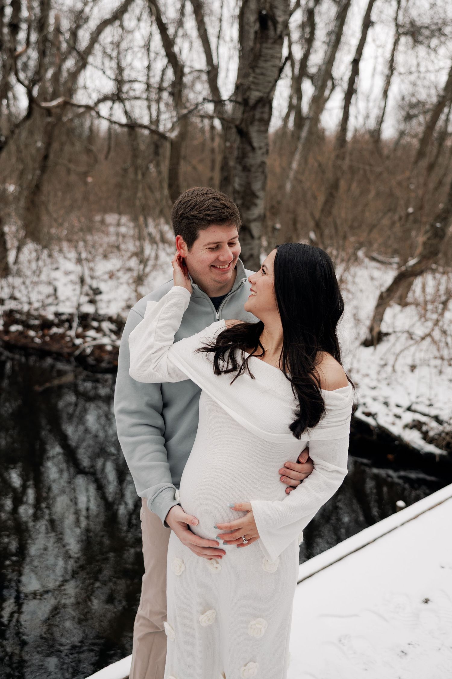 Winter maternity photoshoot of couple embracing on wooden bridge over stream.