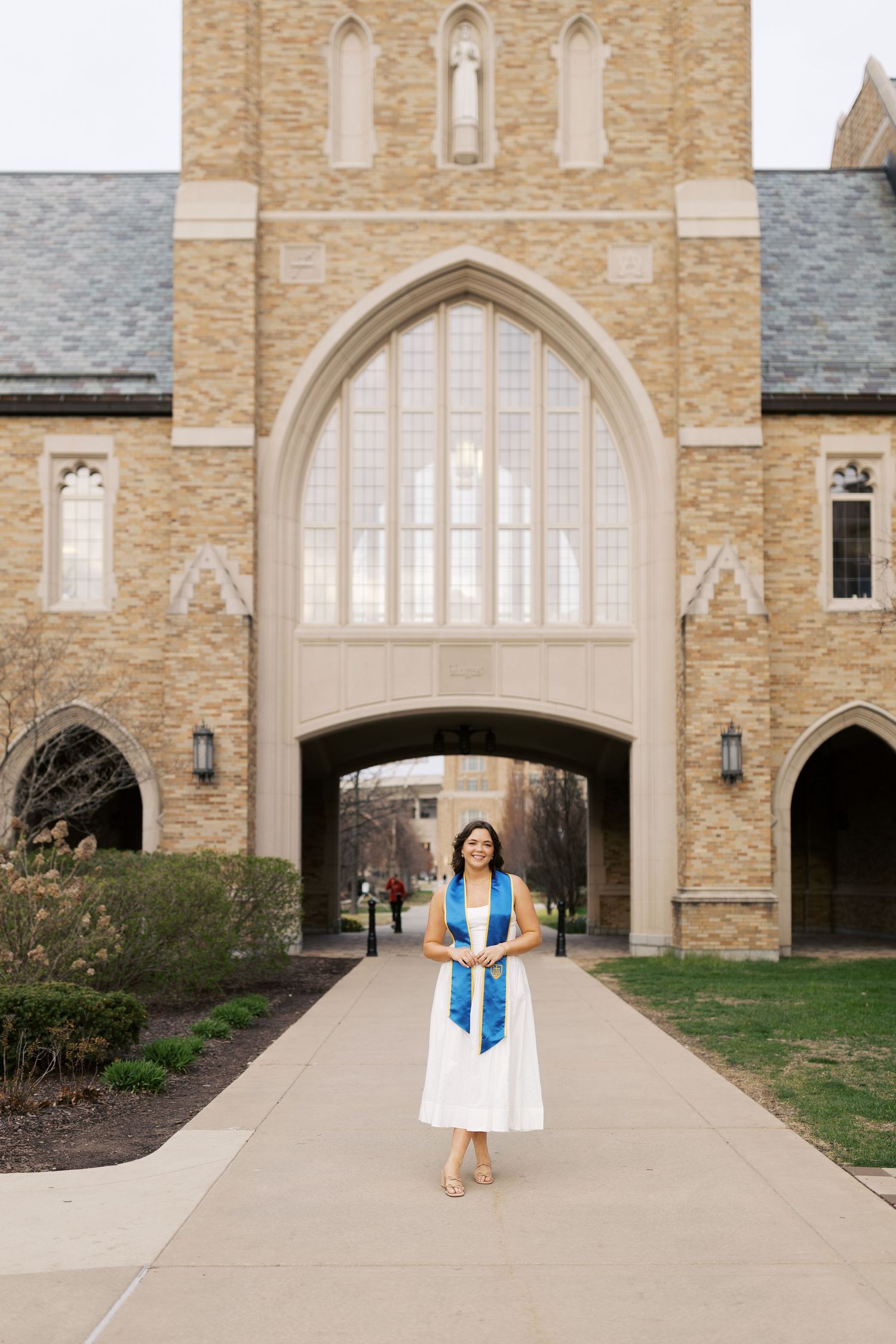 Graduate wearing white dress and blue academic stole stands in front of grand gothic church archway on college campus.