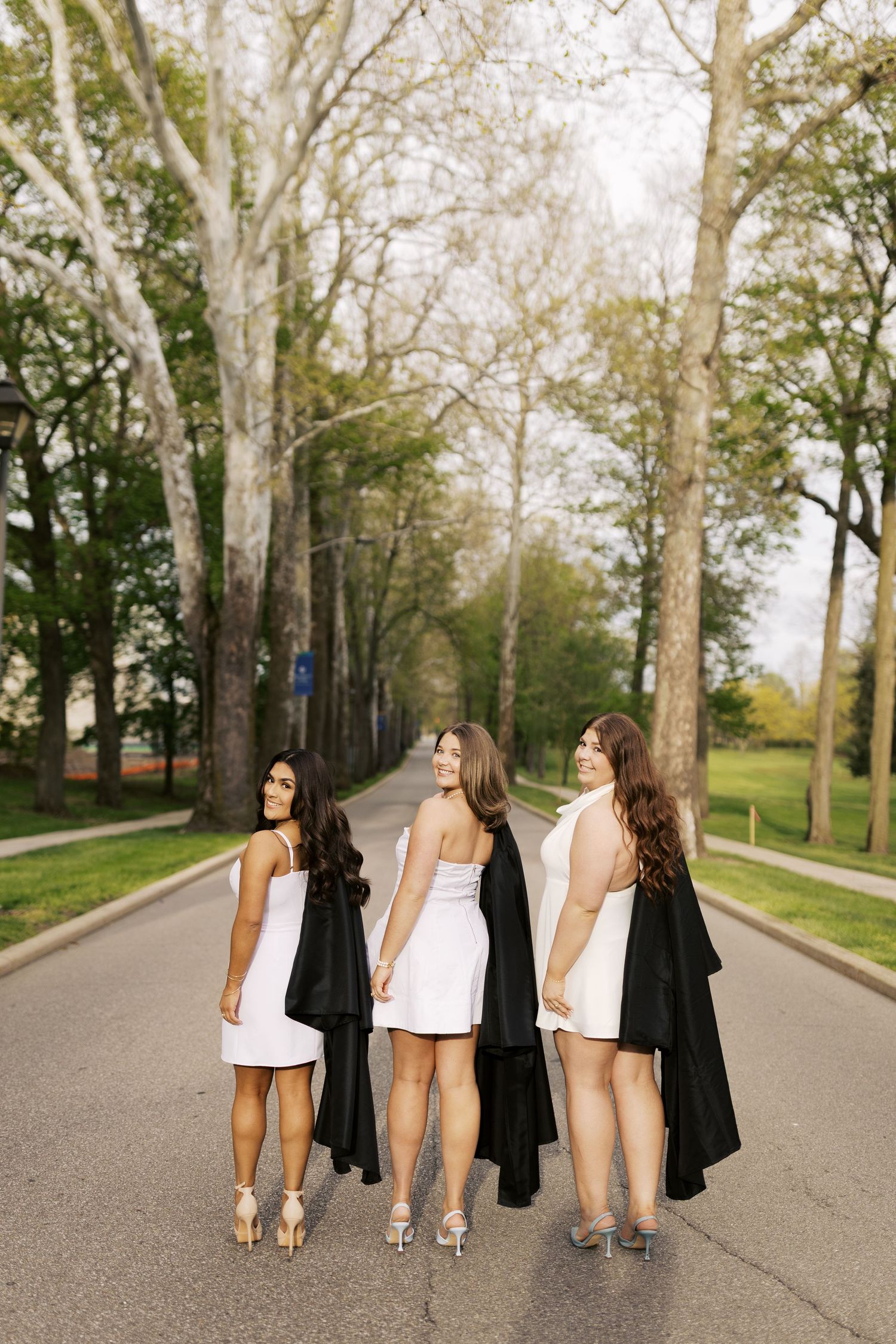 Group walks down tree-lined campus path wearing graduation attire and carrying jackets.
