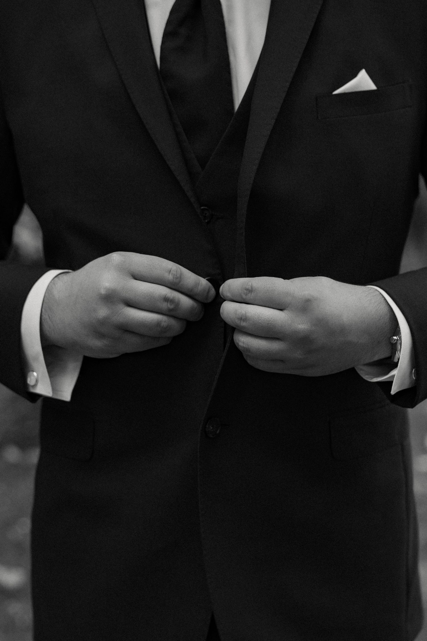 Close-up black and white shot of groom adjusting his suit buttons with crisp white cuffs and pocket square detail.
