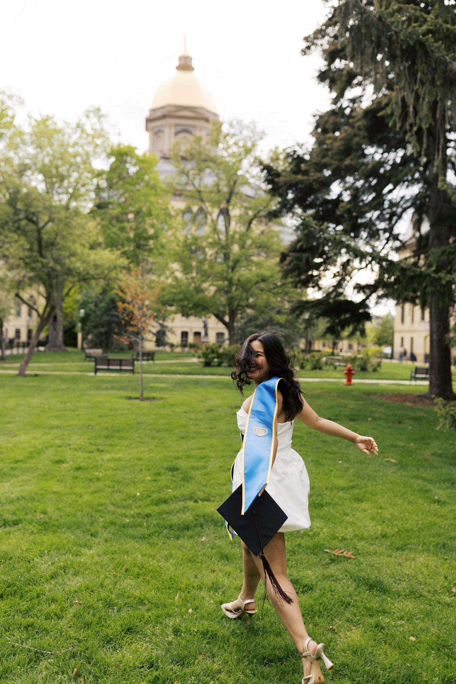Graduate dances joyfully on green lawn with historic campus building dome in background.