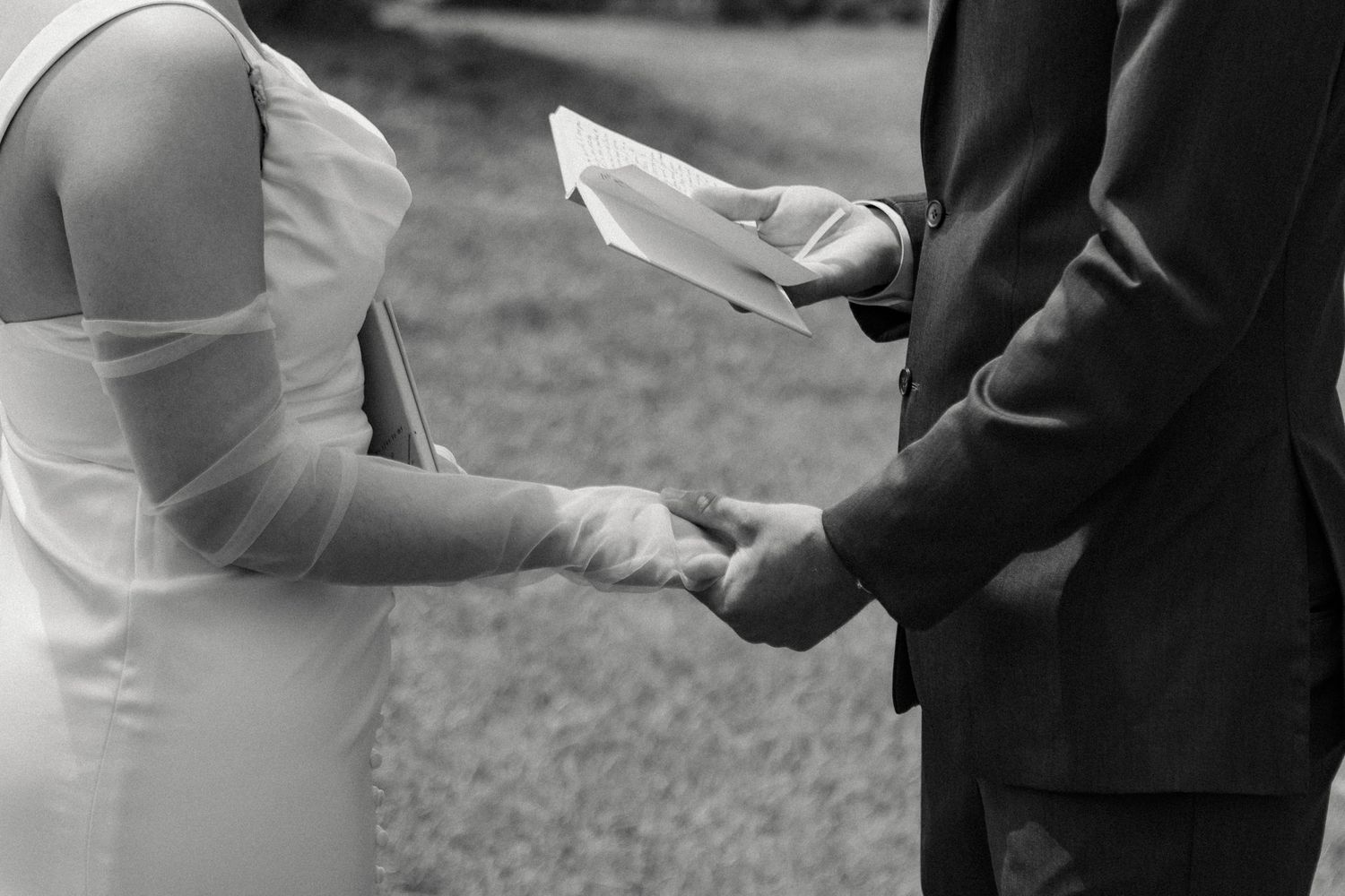 Close-up black and white shot of hands exchanging vows during an outdoor wedding ceremony.