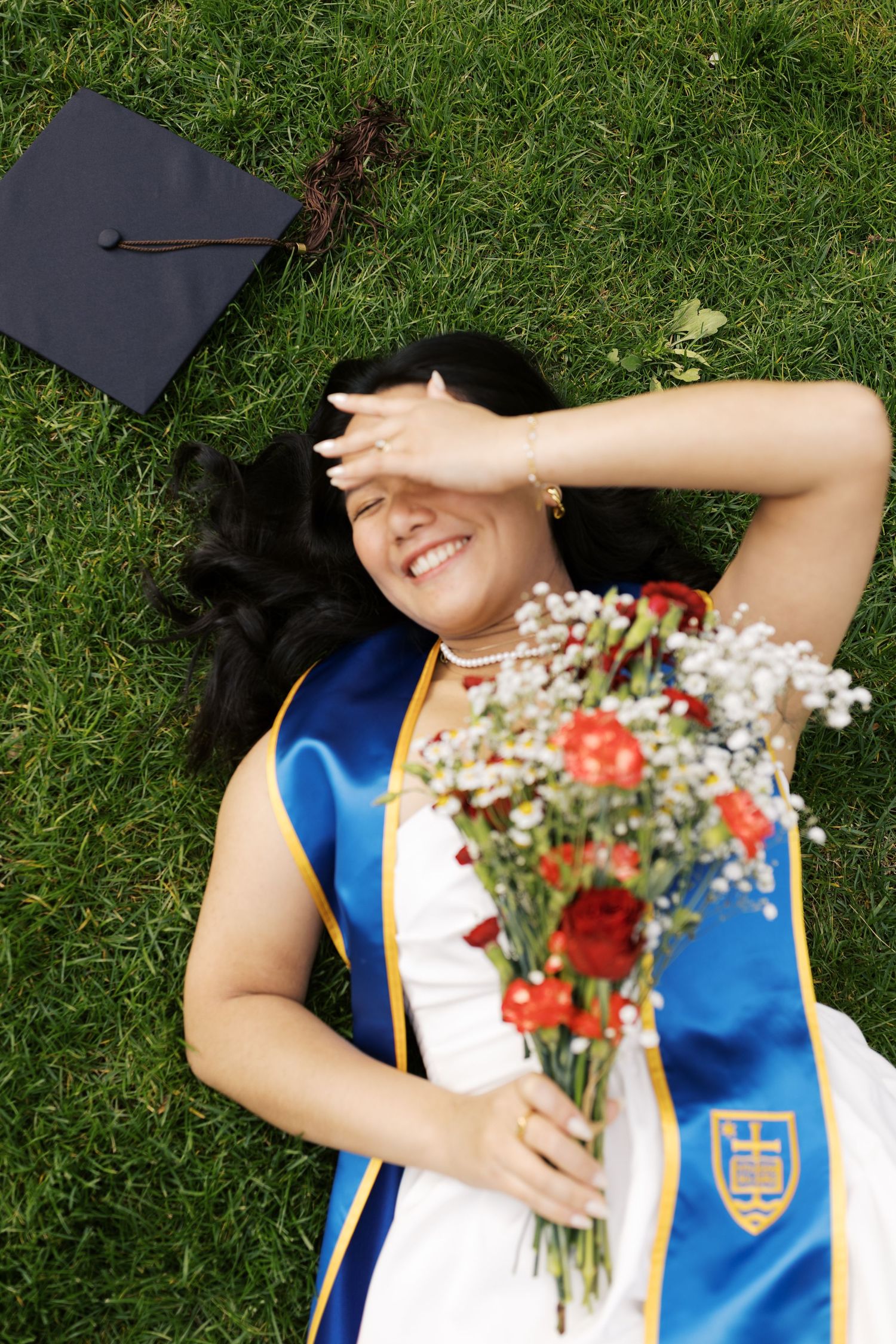 Graduate lying on grass holding bouquet and wearing blue sash with graduation cap.