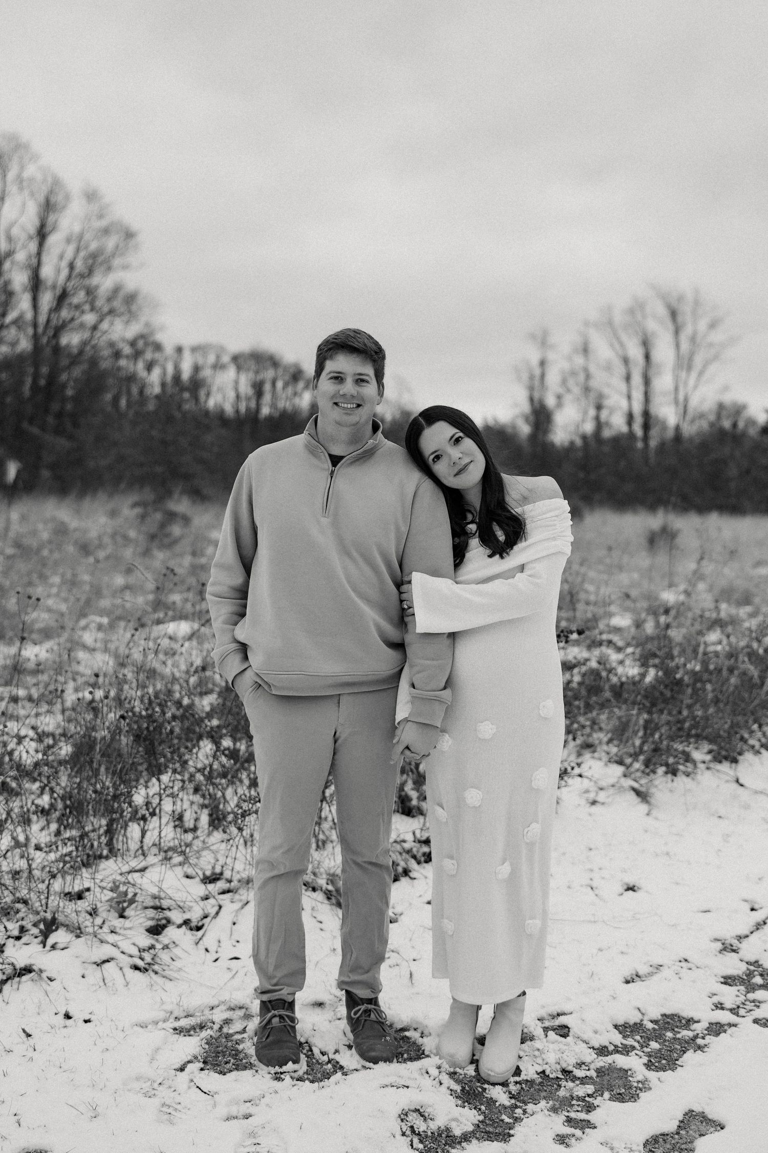 A romantic black and white portrait of a couple standing in a snowy field with bare trees in the background.