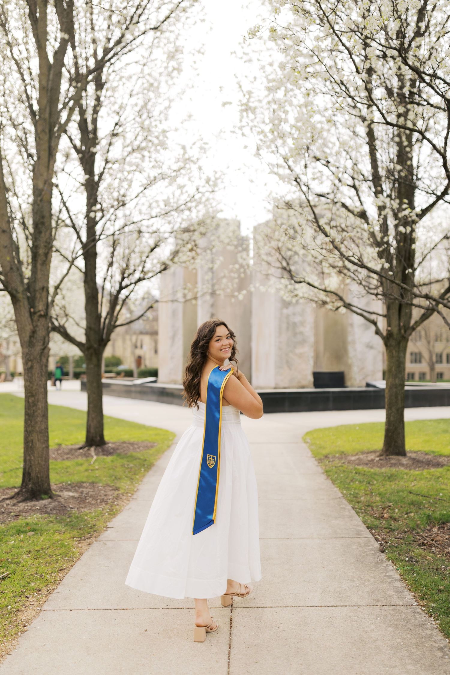 A graduating student in a white dress and blue sash poses among leafless trees along a campus walkway.