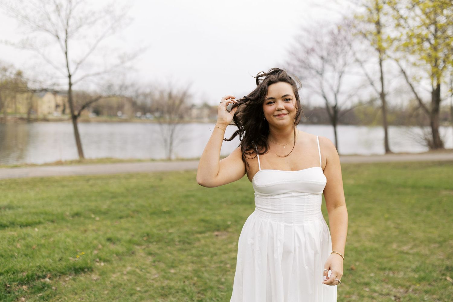 A person wearing a white sundress stands in a grassy park area with a lake in the background on an autumn day.