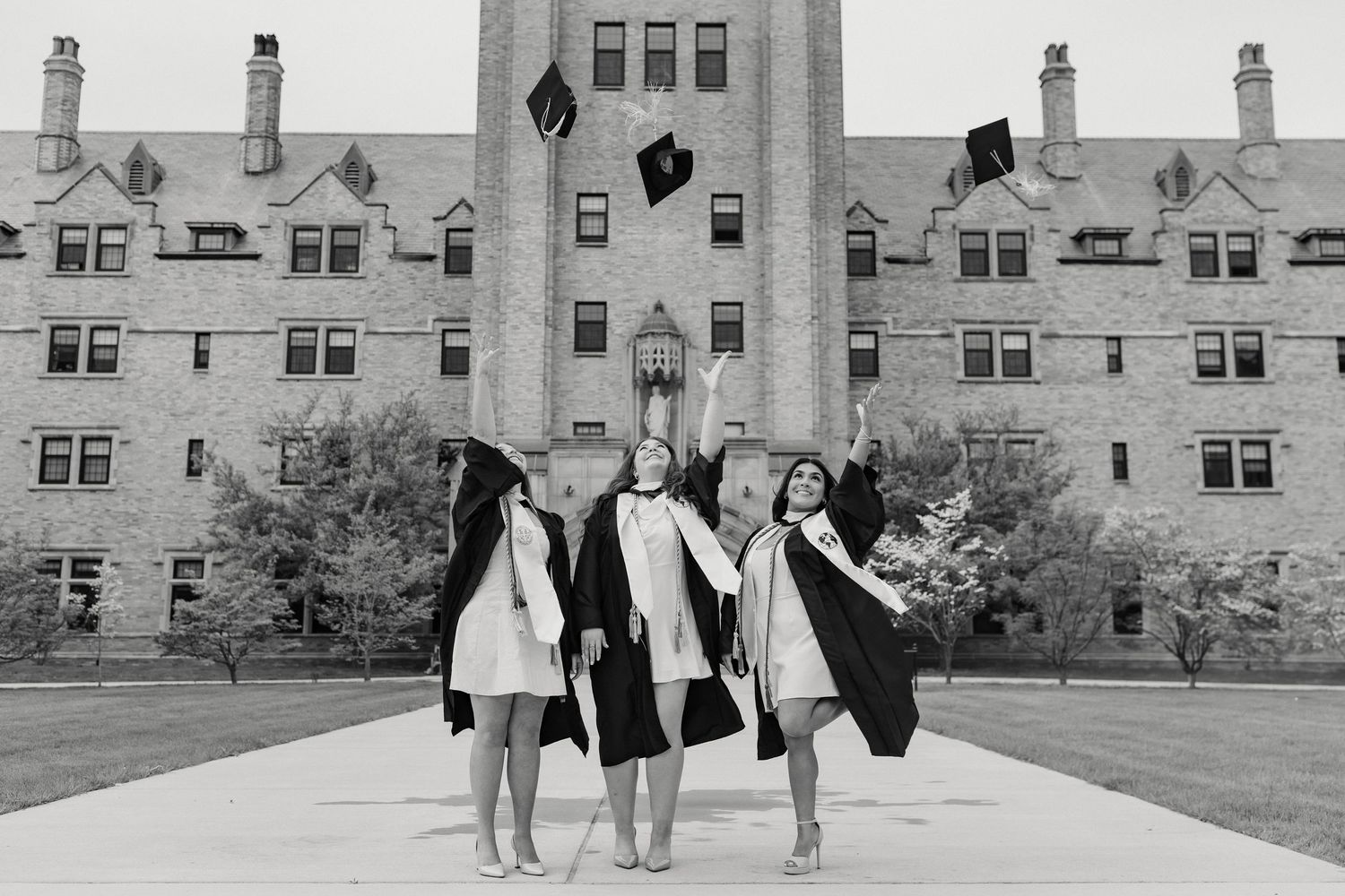 A black and white graduation photo of three graduates celebrating on campus.