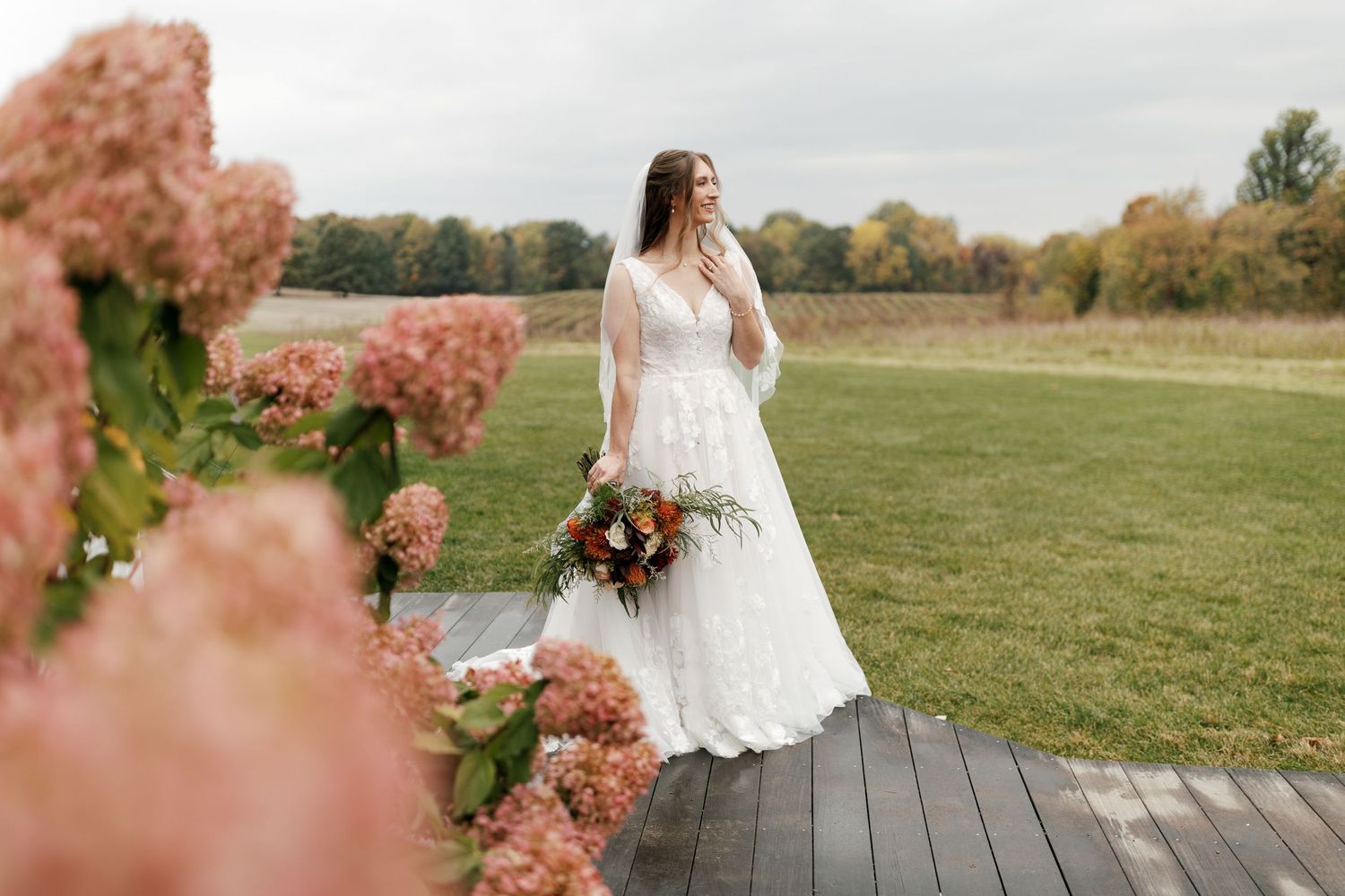 Bride in white lace gown holding bouquet stands on deck surrounded by pink hydrangeas in lush green field.