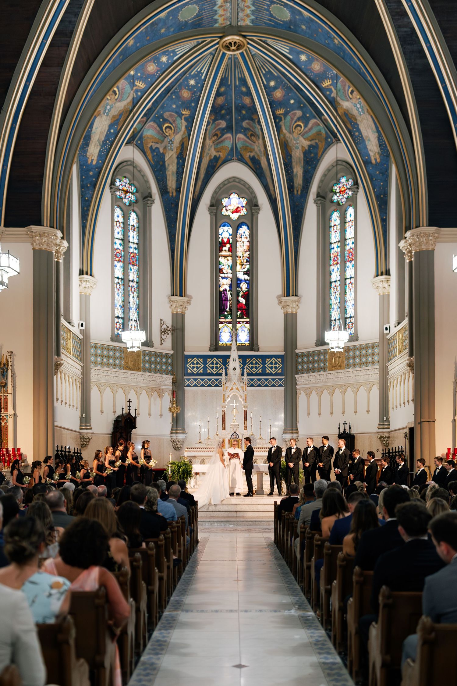 A wedding ceremony in a grand cathedral with blue and gold vaulted ceilings and stained glass windows.