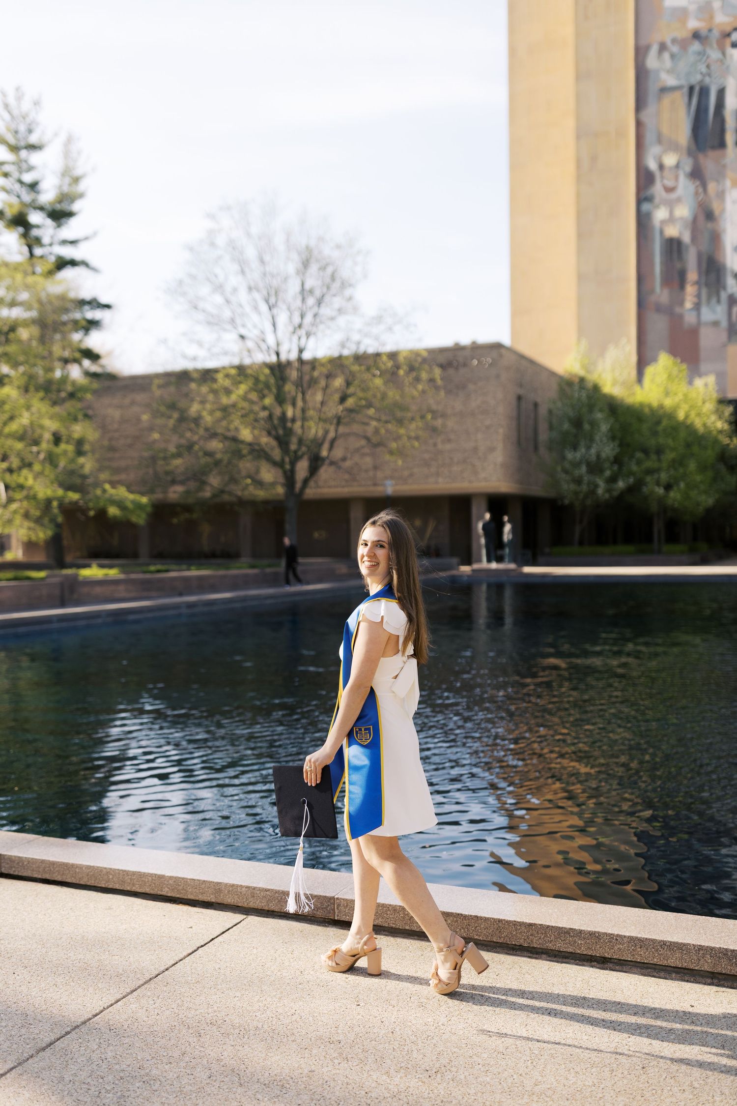 Graduate walks along reflective pool outside historic university building.