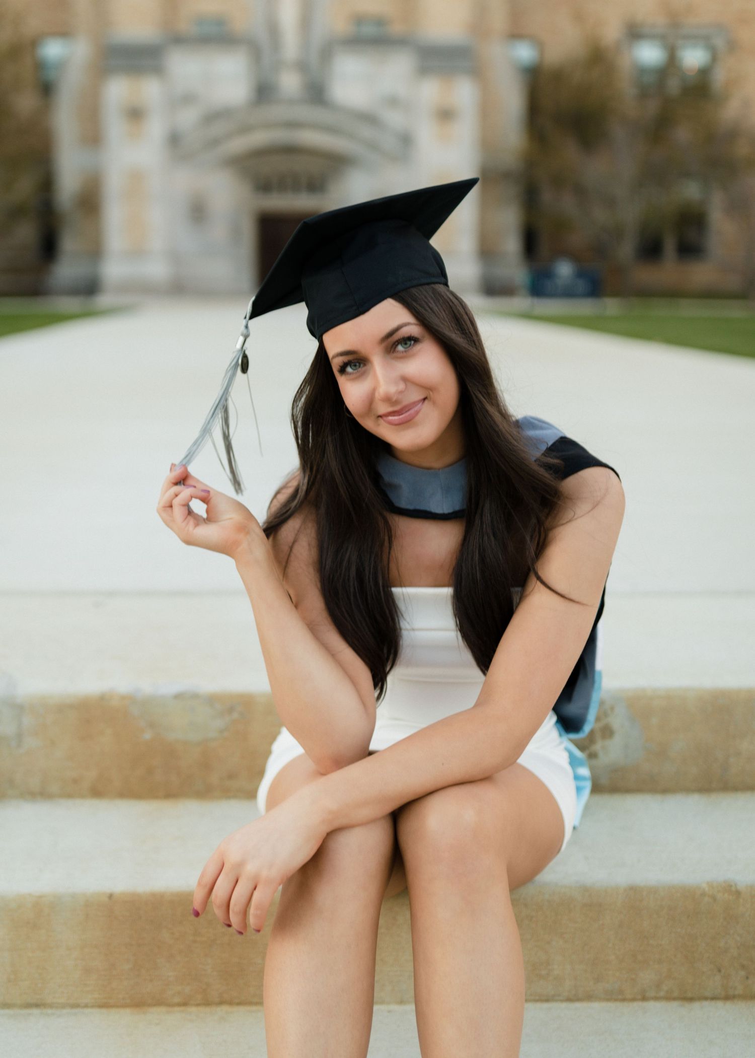 Graduate in cap and gown sits on steps holding tassel while posing in front of campus building.