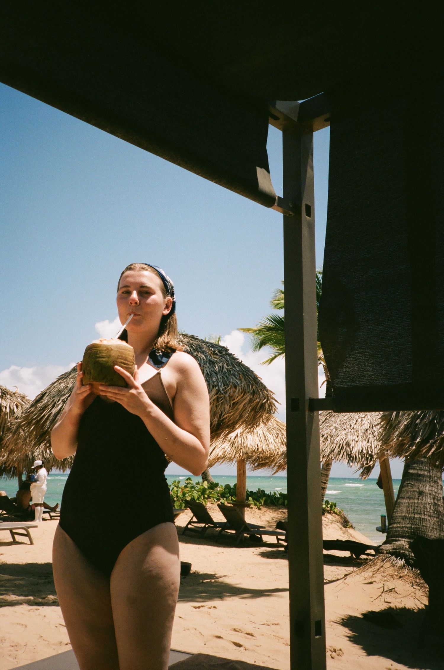 A person relaxing under a beach cabana with thatched umbrellas visible in the background.