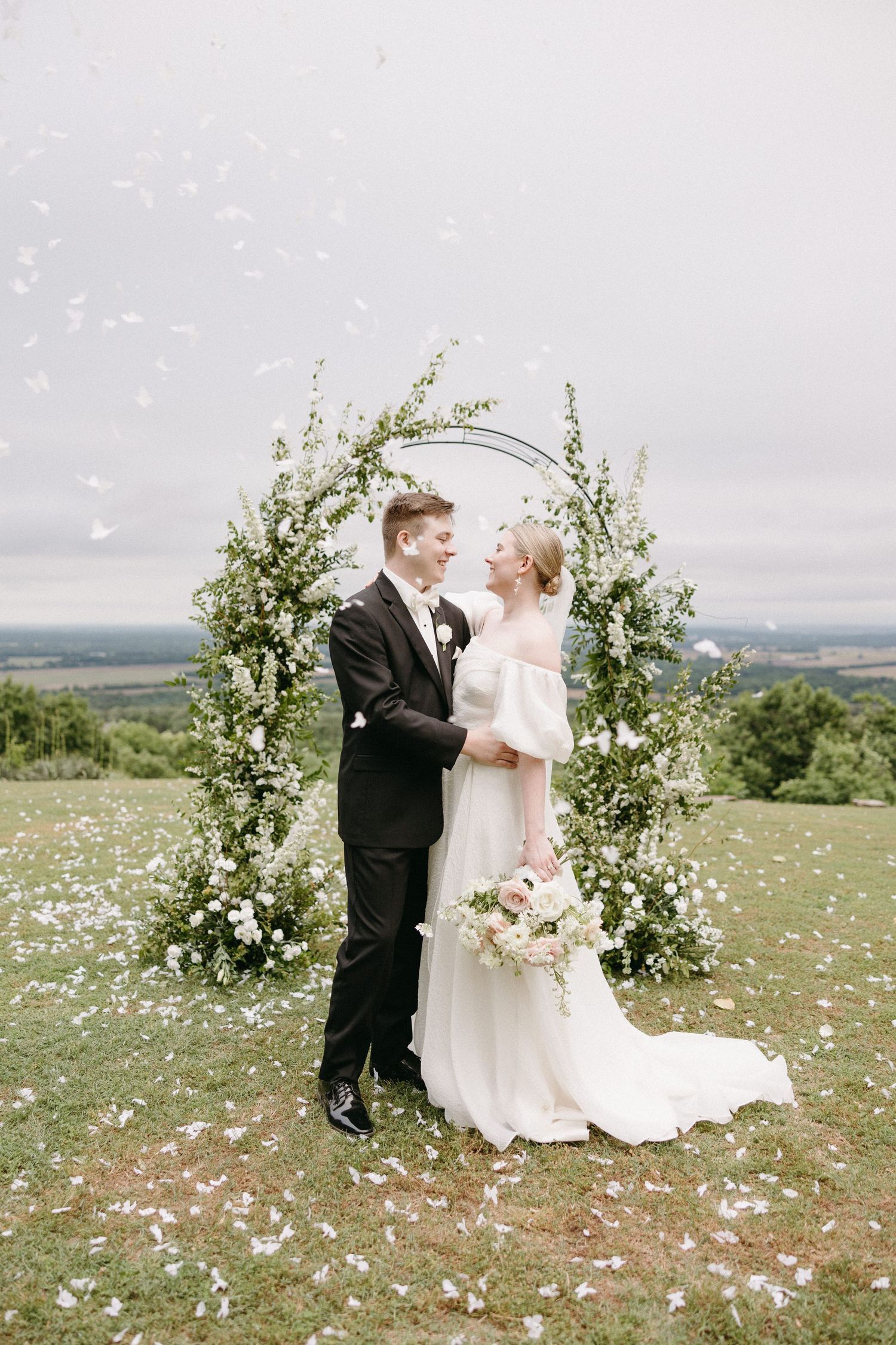 Wedding couple poses in a scenic outdoor setting with floral arch and scattered petals in a meadow.