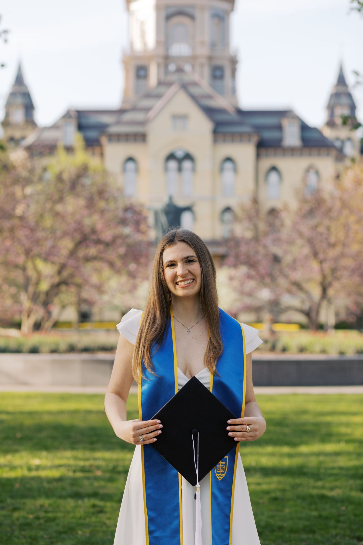 Graduate poses in front of historic Gothic university building wearing blue and white graduation regalia.