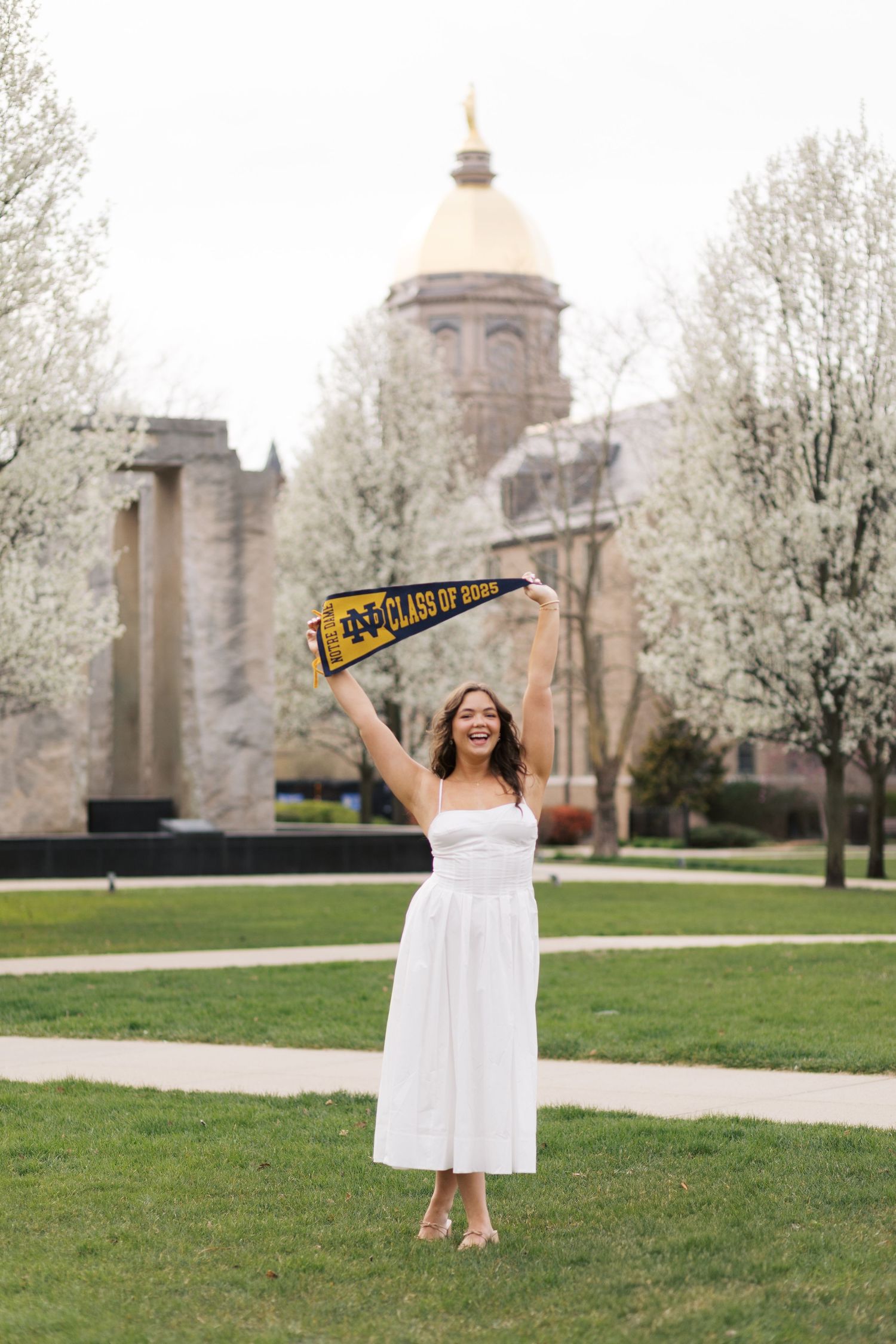 Graduate in white dress celebrates on university campus with raised pennant flag near historic domed building.