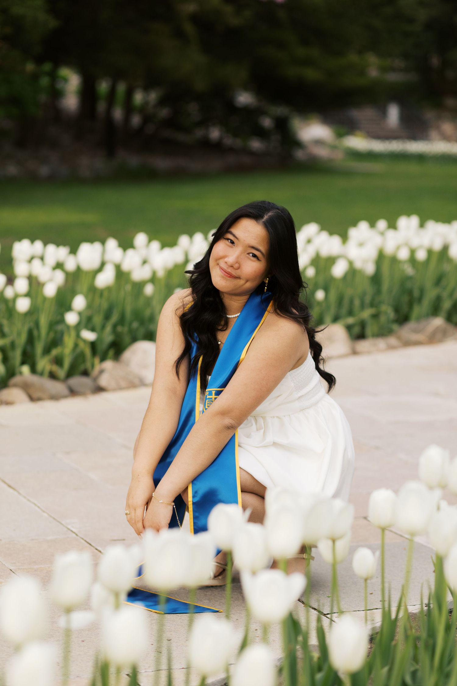 Graduate sits among white tulip garden wearing graduation sash and white dress.