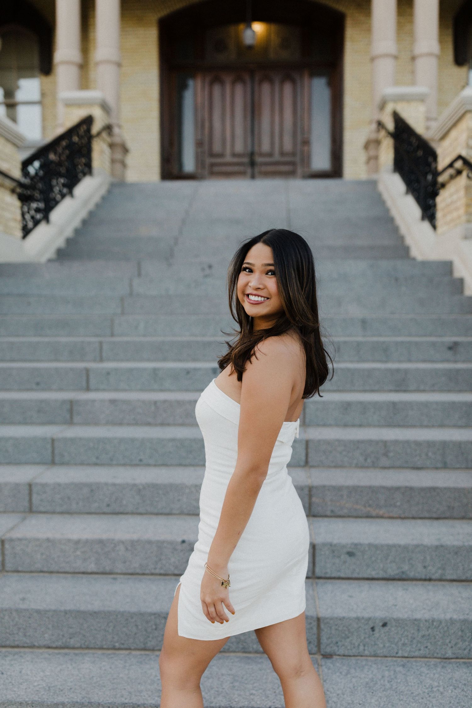 Woman in white dress poses on grand stone steps of historic building.