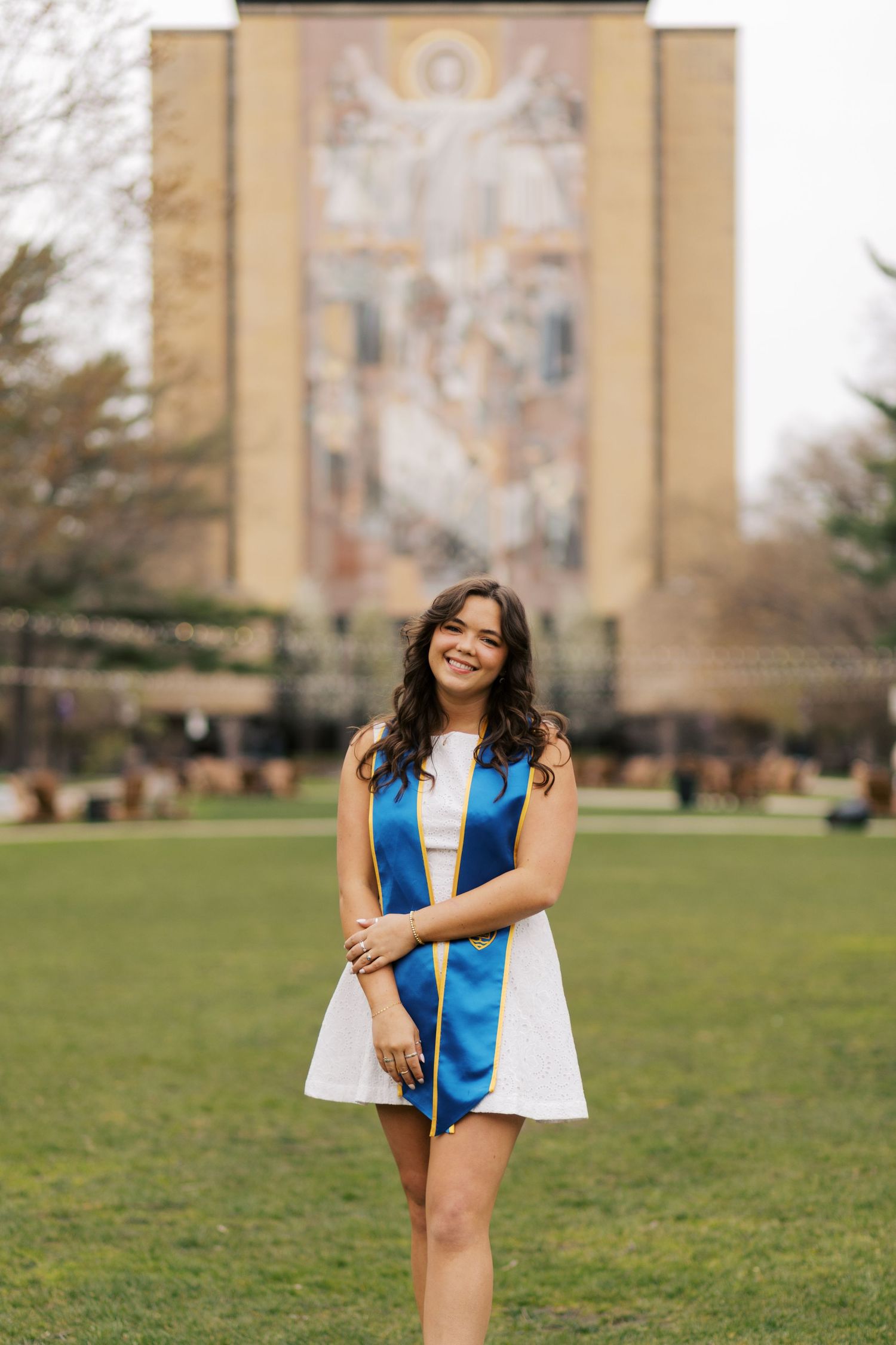 A graduate stands on a grassy lawn in front of a university building wearing academic regalia with a blue stole.