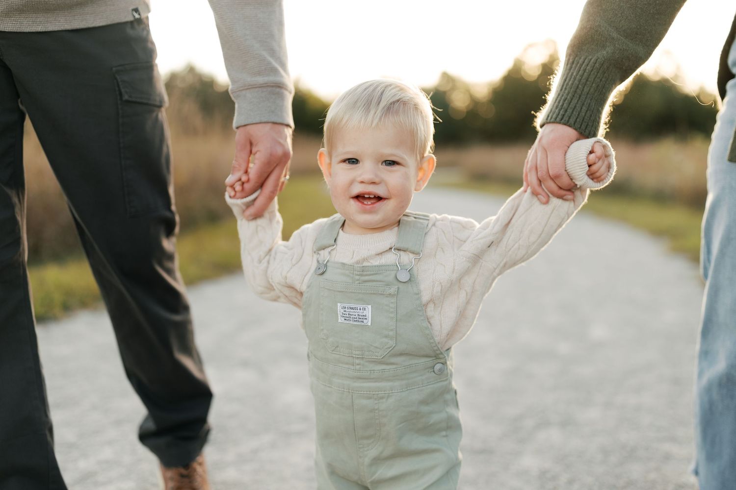 A toddler in overalls smiling while holding hands with parents on a path.