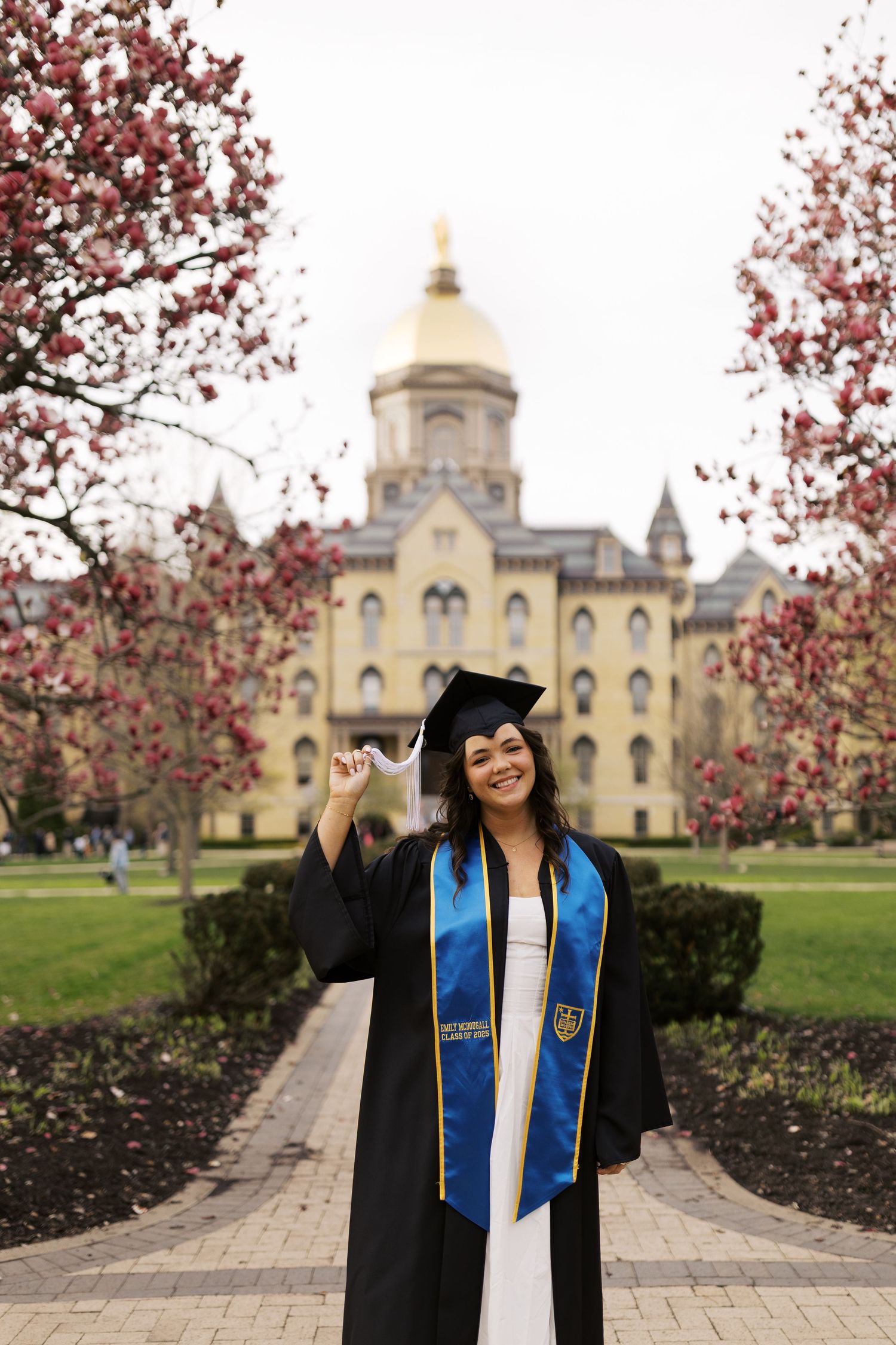 Graduate in cap and gown poses in front of Notre Dame's Golden Dome building with spring cherry blossoms in bloom.
