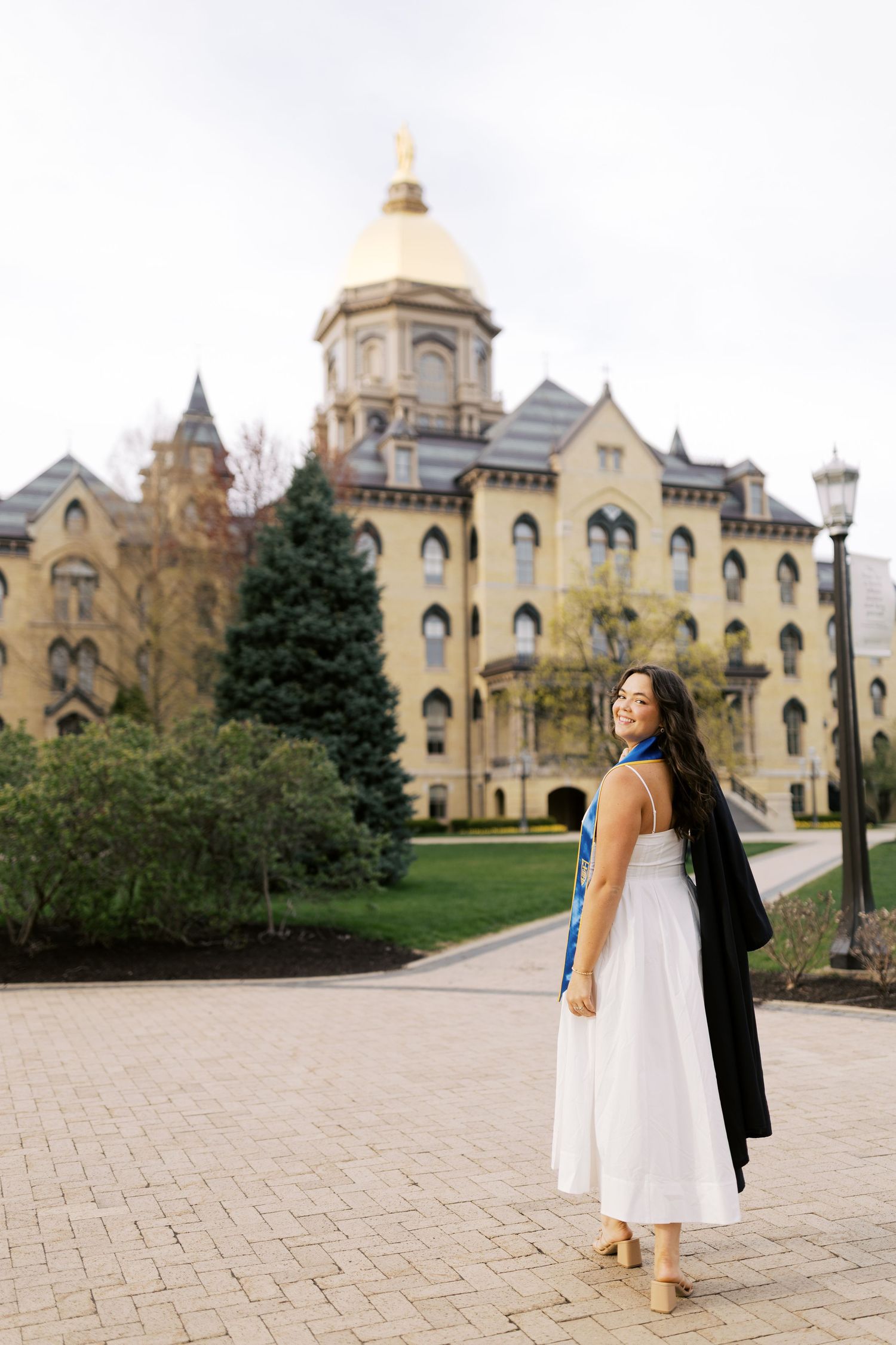 A person in a white dress stands along a brick path in front of a historic Gothic Revival university building.