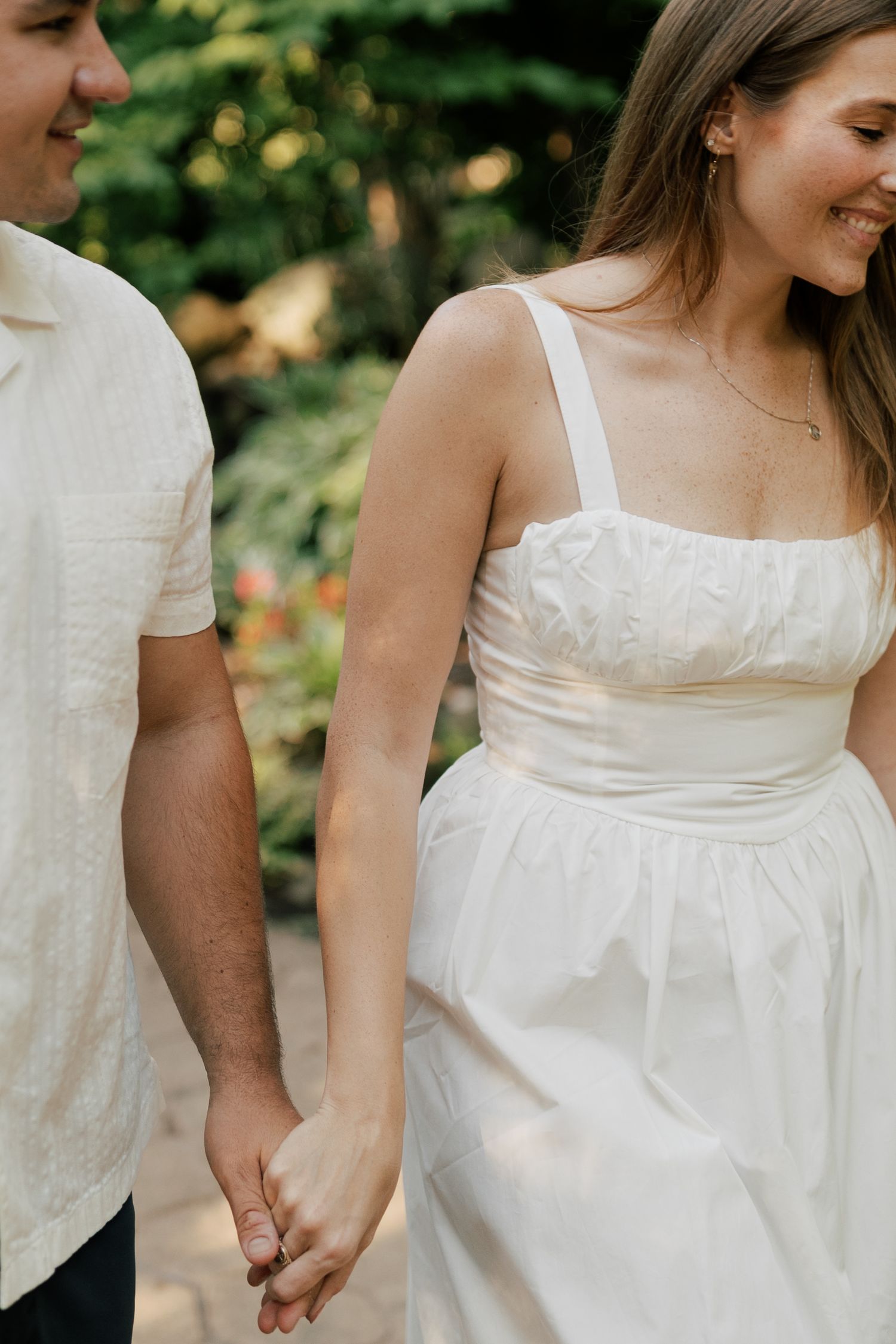 A couple holding hands outdoors, wearing white clothing and sharing a joyful moment in a garden setting.