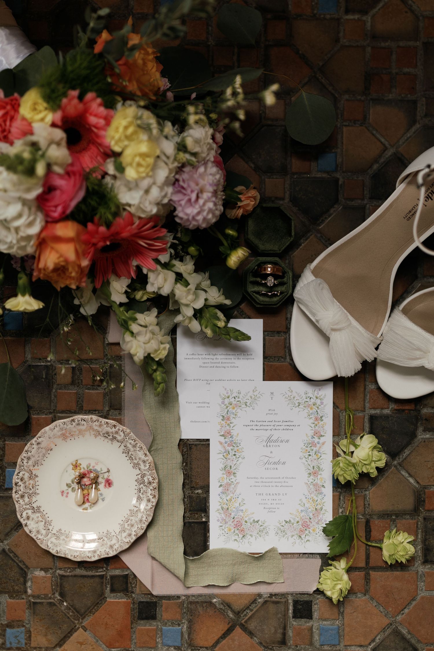 Wedding details flatlay with floral invitation, vintage plate, bridal shoes and colorful bouquet on tiled floor.