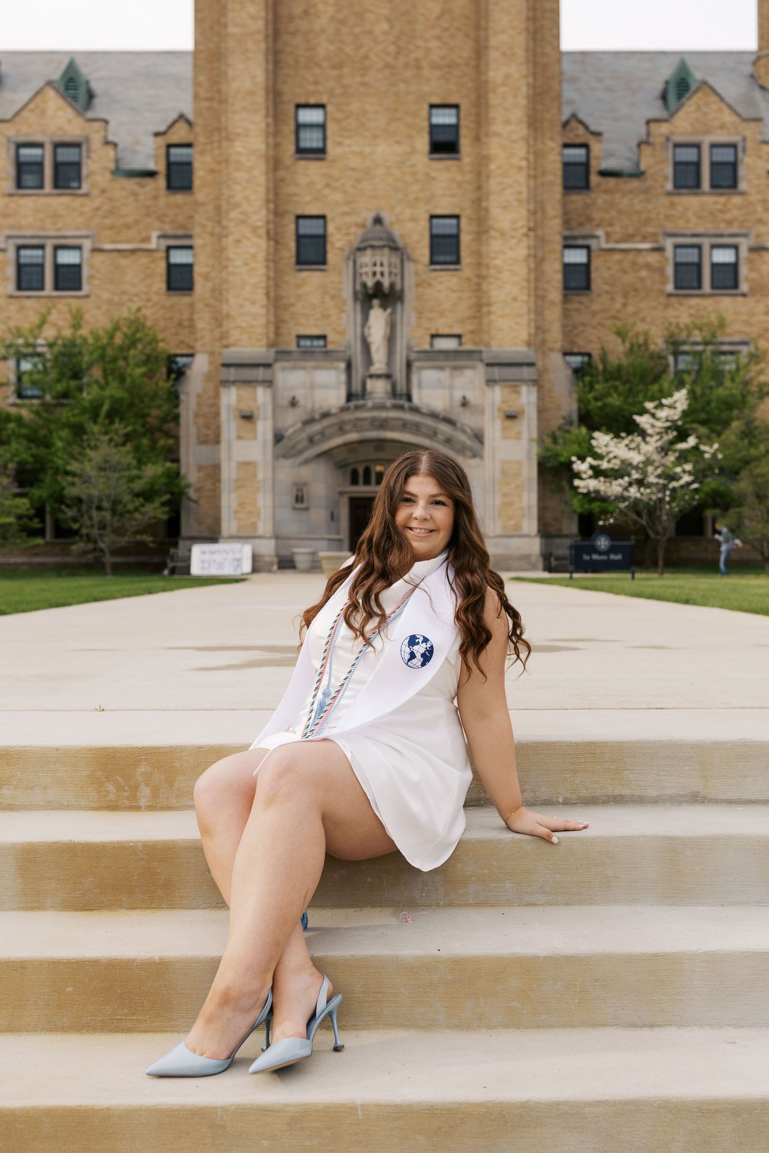Graduate poses on stone steps in front of historic university building wearing white dress and graduation sash.