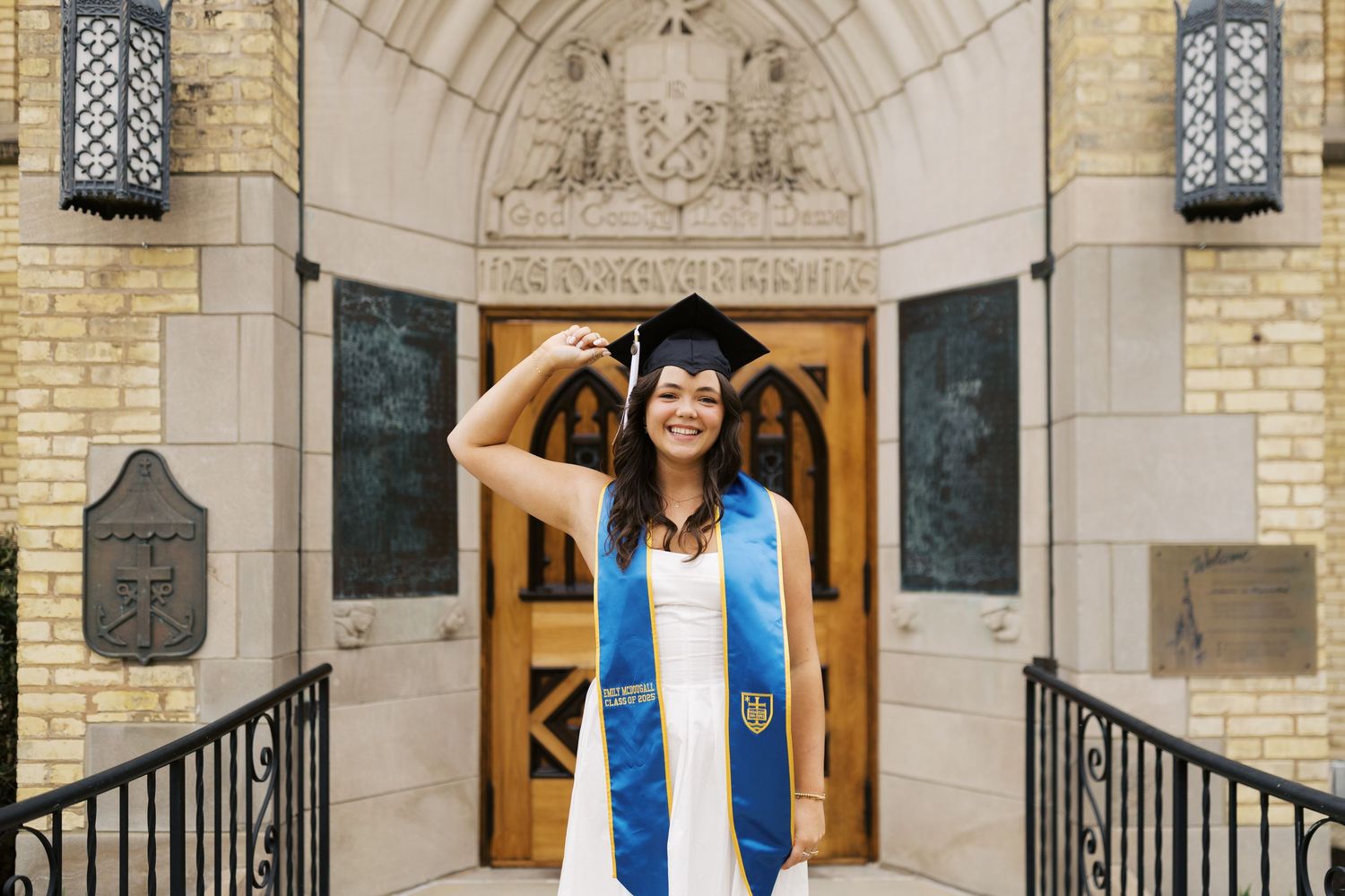 Graduate in blue and white regalia stands in front of ornate stone building entrance with arched doorway and railings.