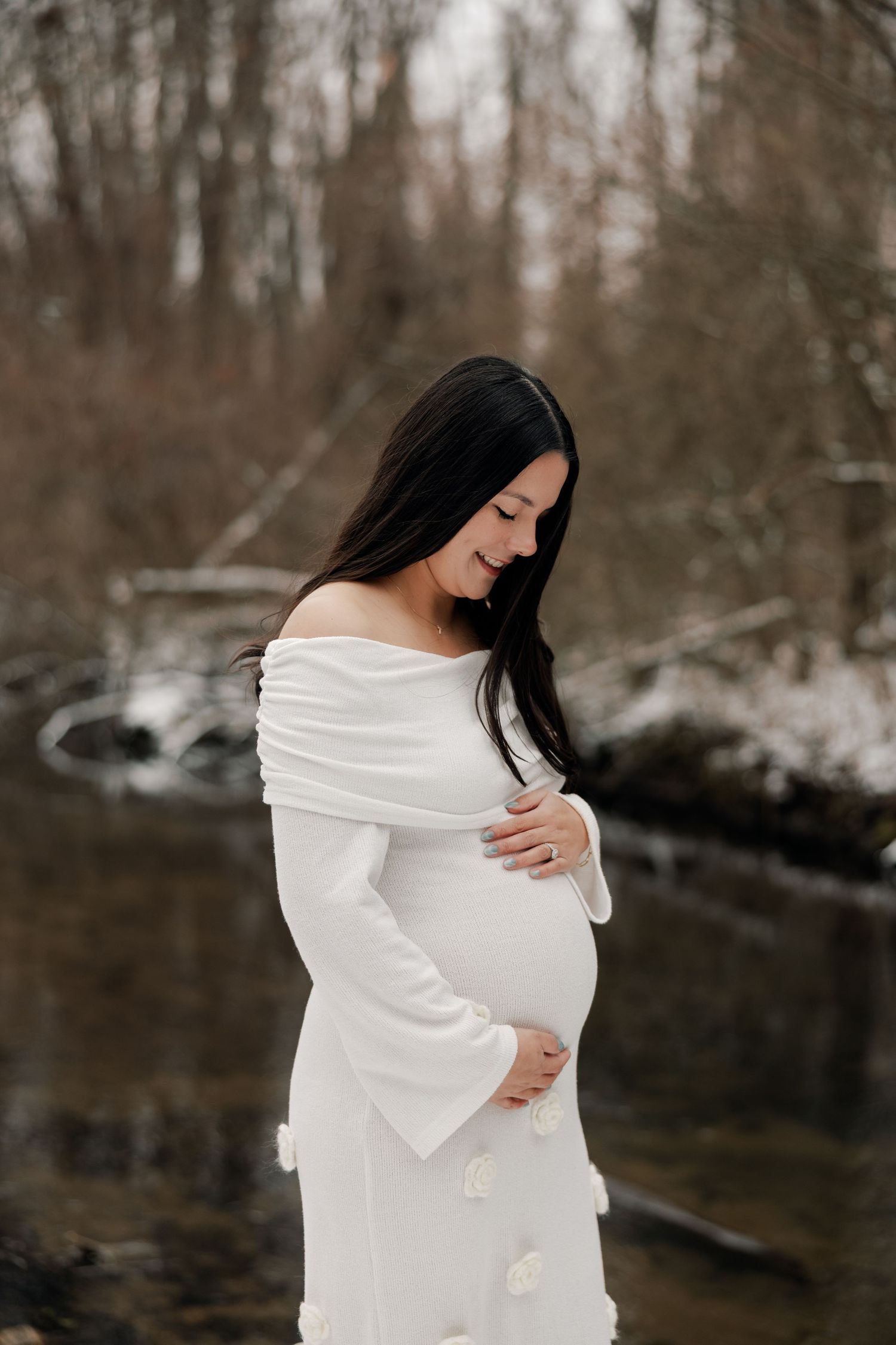 Maternity photo of woman in white dress standing by snowy stream.